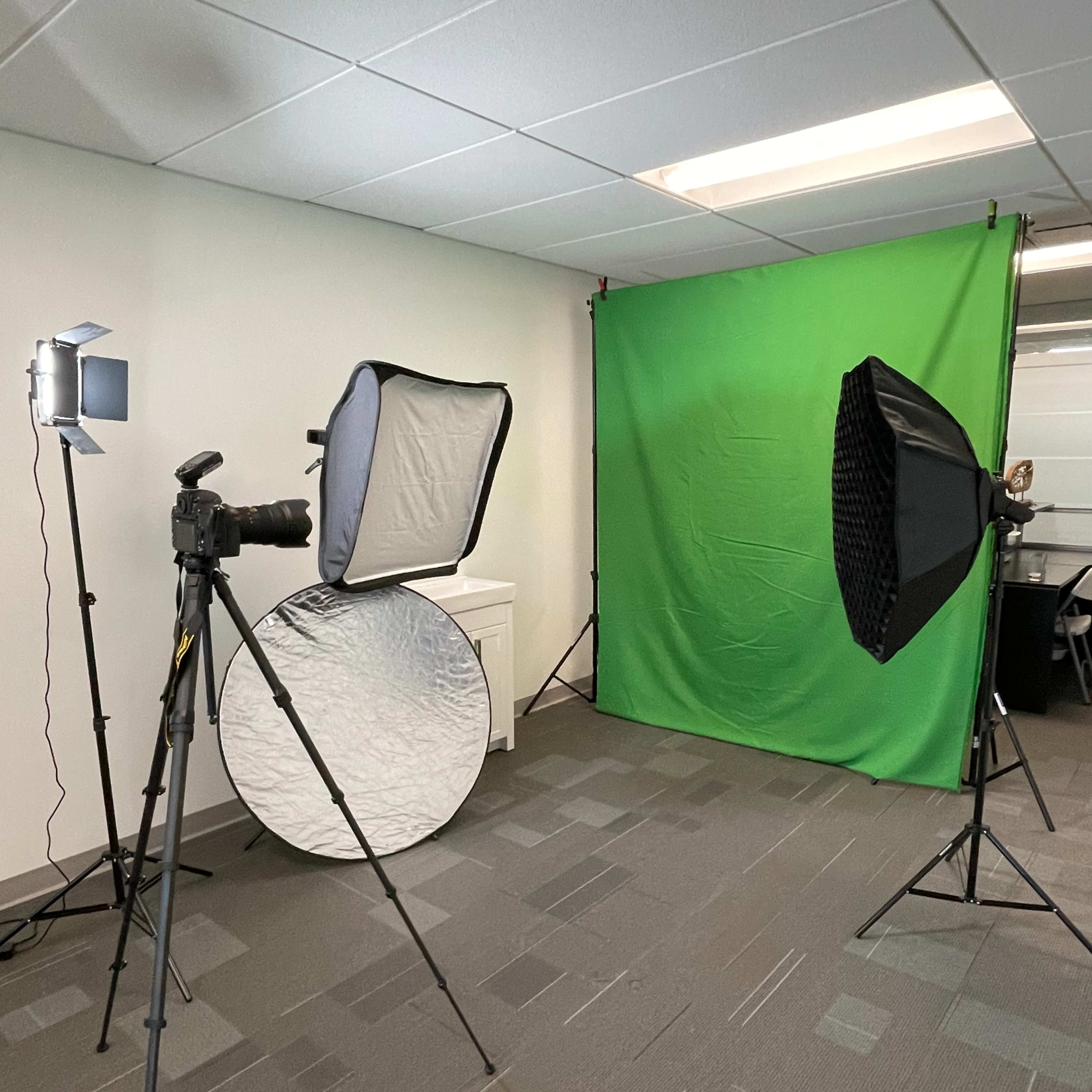 A photography setup featuring a green screen, softbox lights, a camera on a tripod, and a reflective disc in a room with gray carpeting.