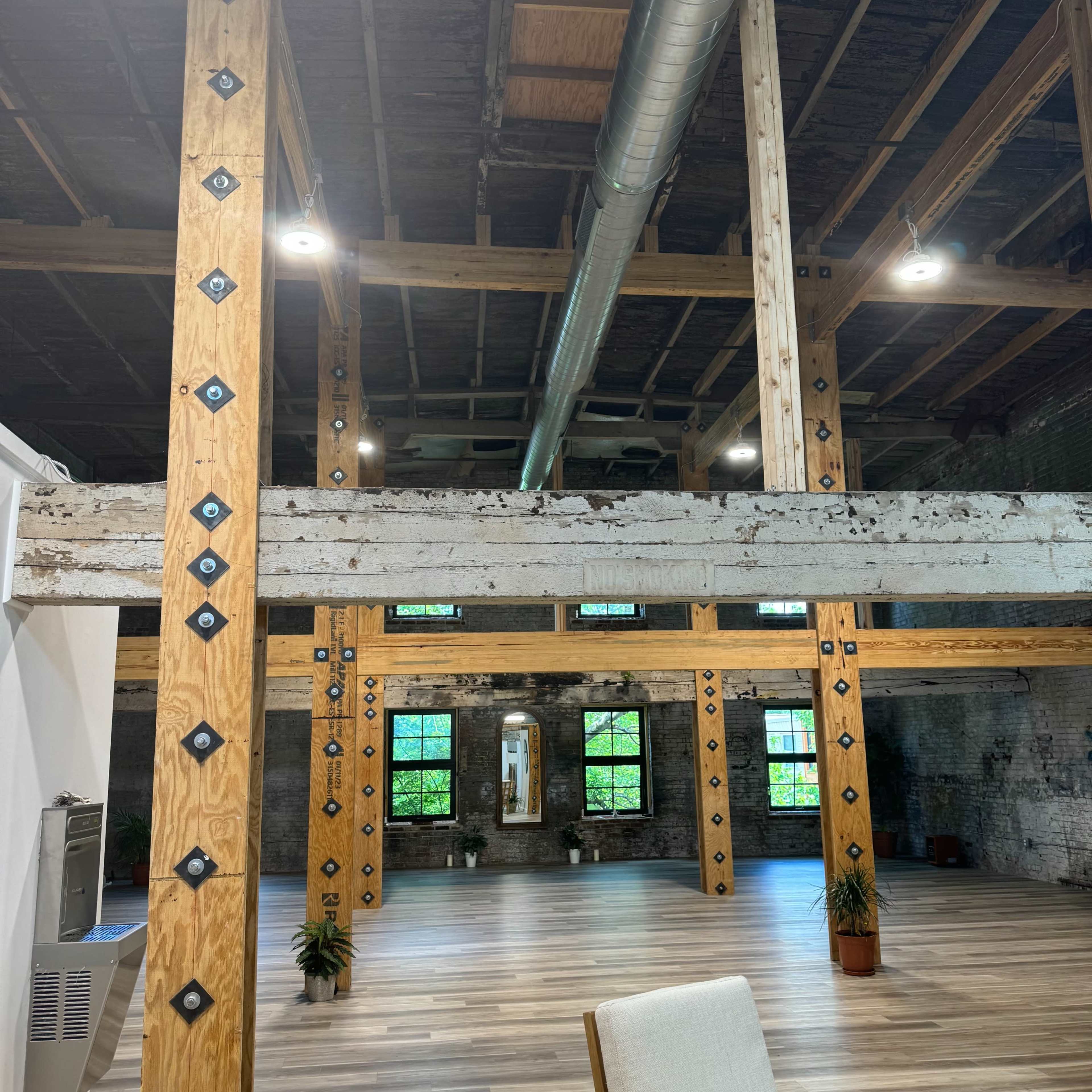 An industrial-style interior space with exposed wooden beams, metal accents, and large windows overlooking a wooden floor.