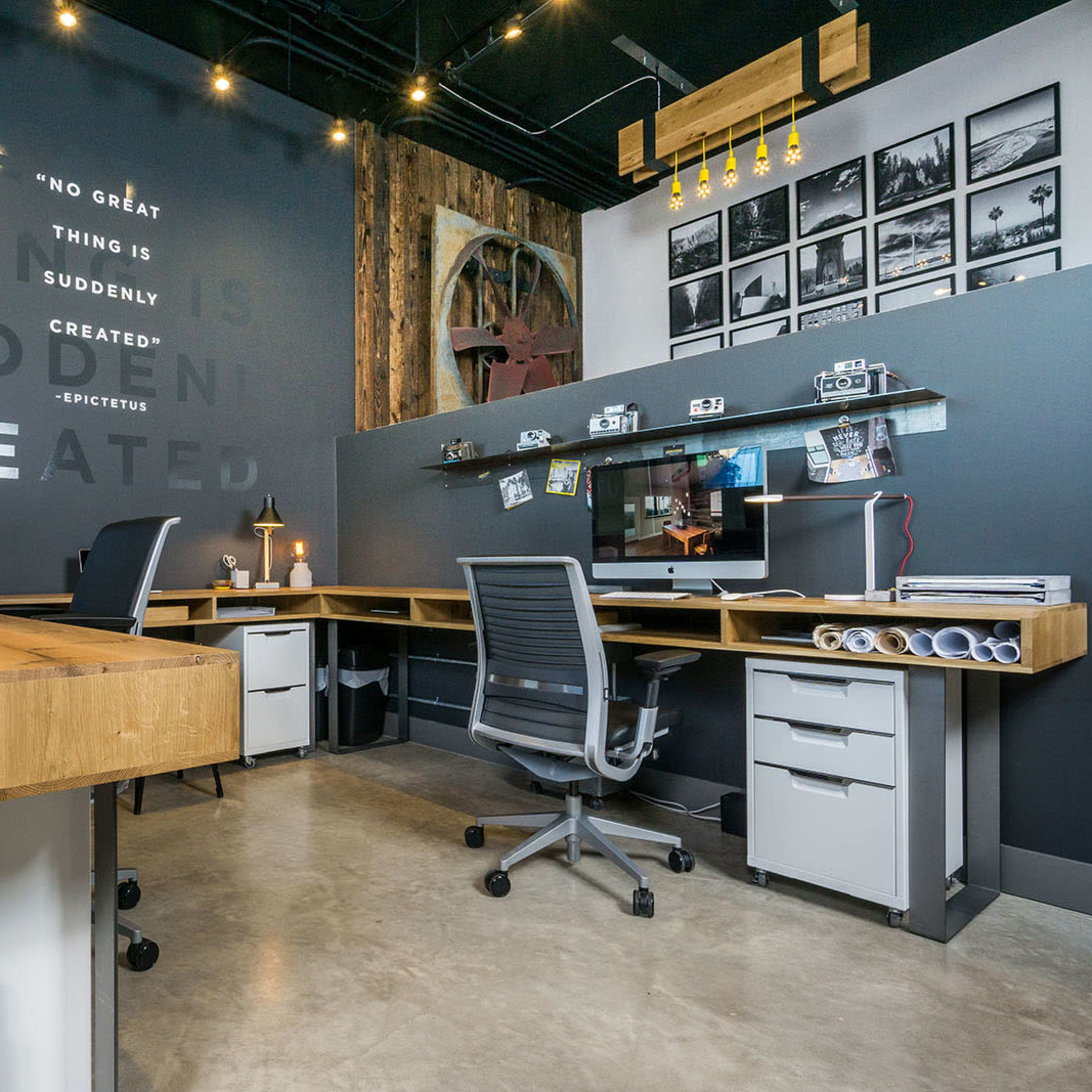 A modern office space with two desk workstations, wooden accents, and a dark wall featuring framed photographs and text.