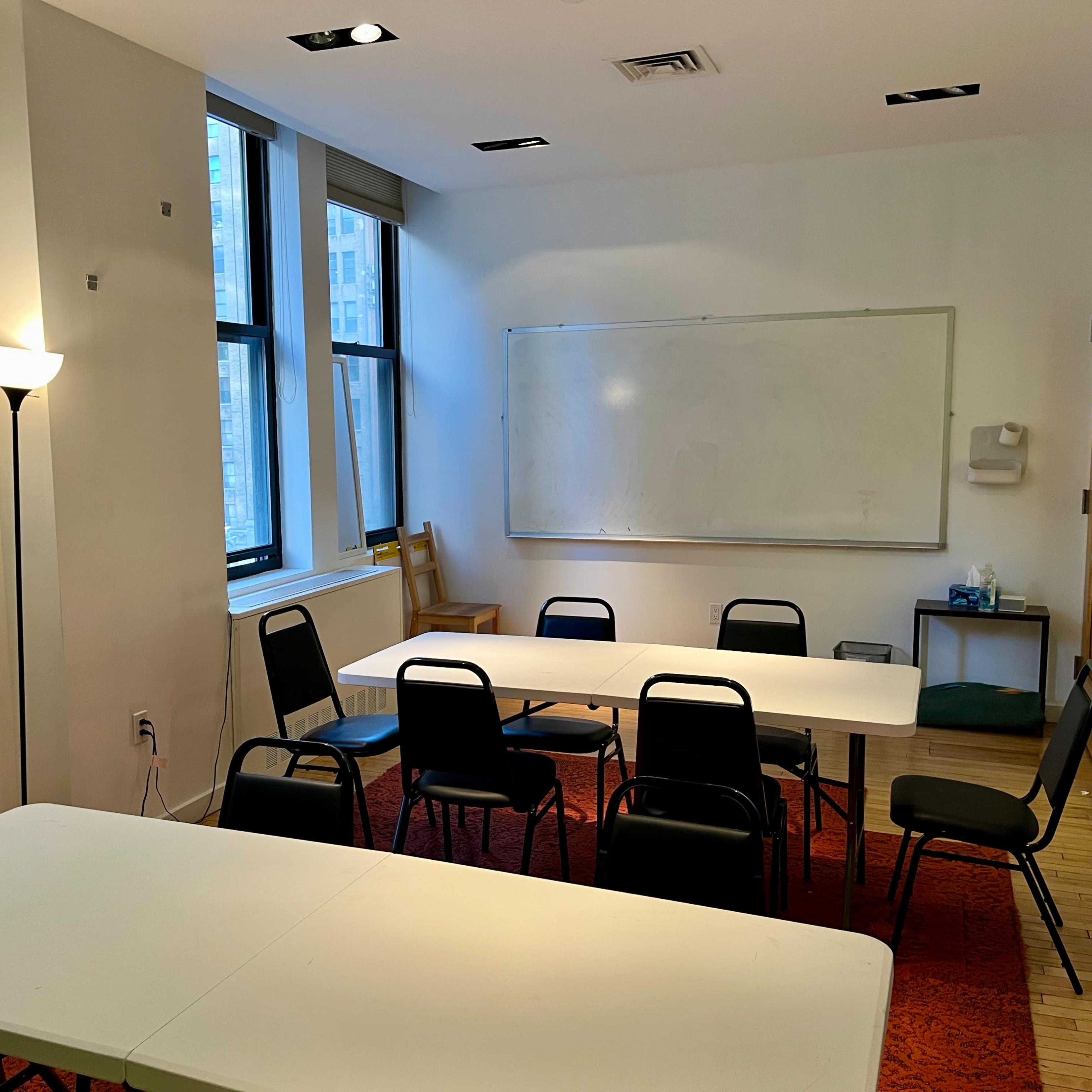 A simple classroom with two tables, several chairs, a whiteboard, and a lamp beside a window.
