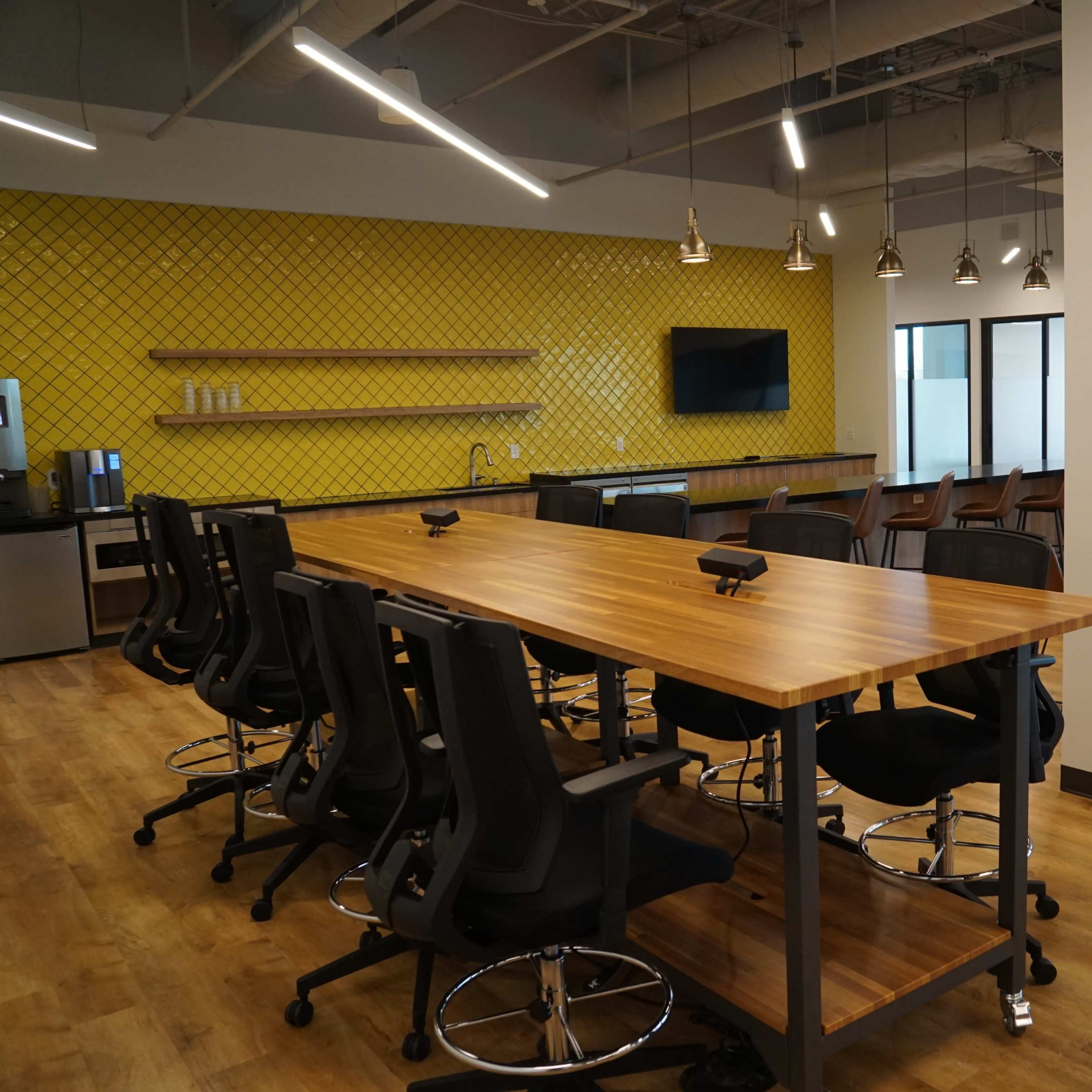 A modern office space featuring a large wooden table with high-backed chairs, a kitchenette area, and several round dining tables against a bright yellow accent wall.
