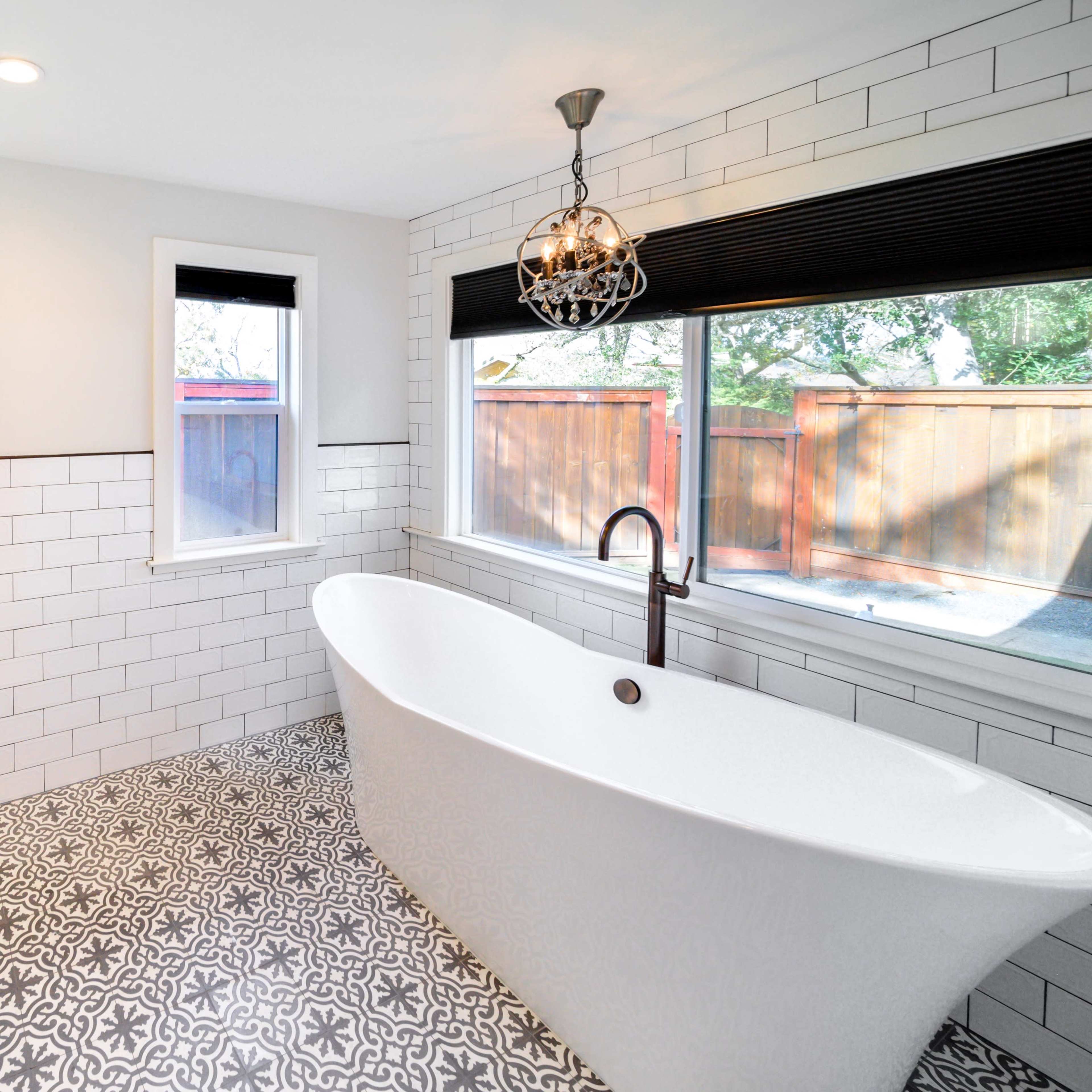 A modern bathroom features a freestanding bathtub, patterned tile flooring, and large windows with wooden blinds.