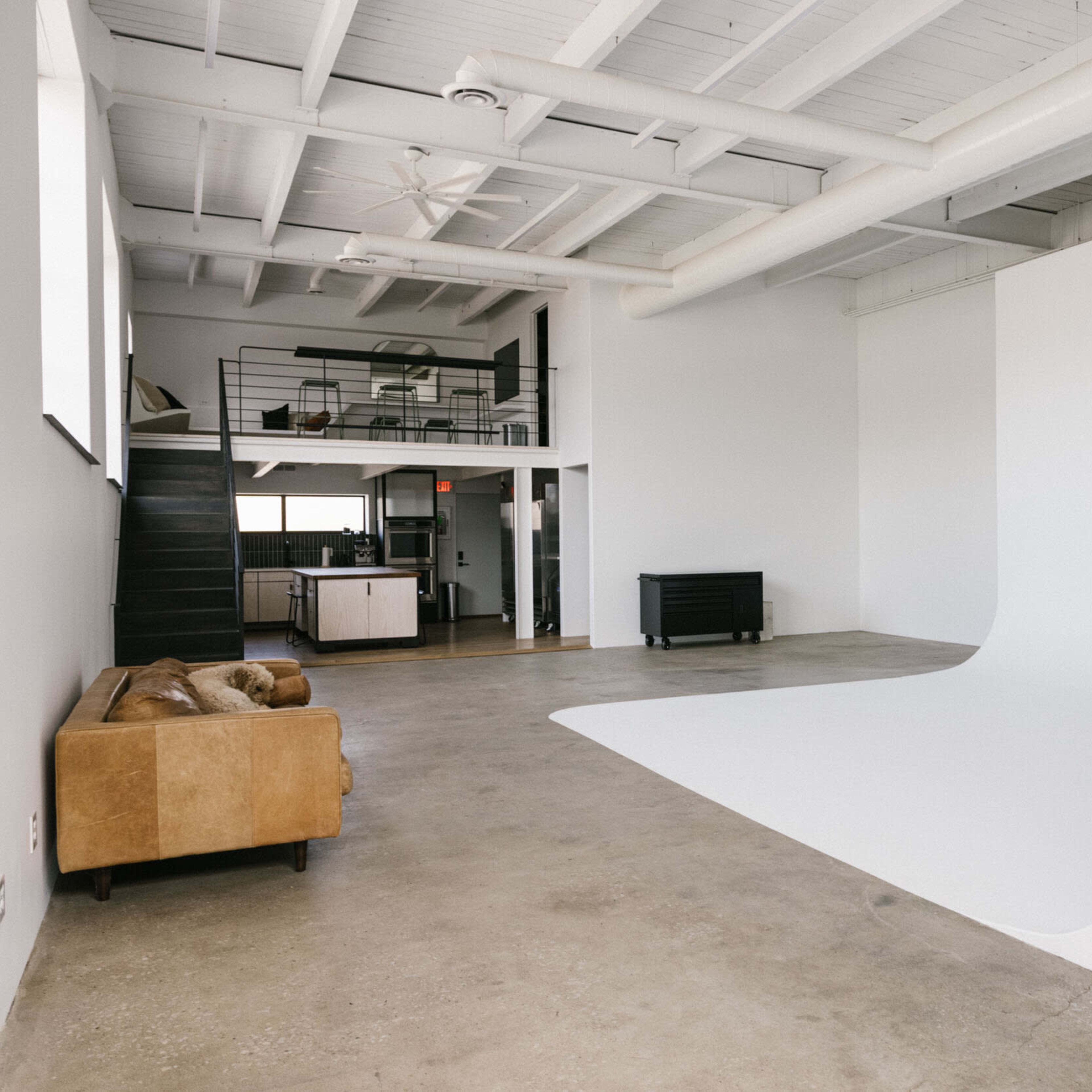 A spacious, minimalistic studio with a curved white backdrop, a brown sofa, and a staircase leading to an upper level.