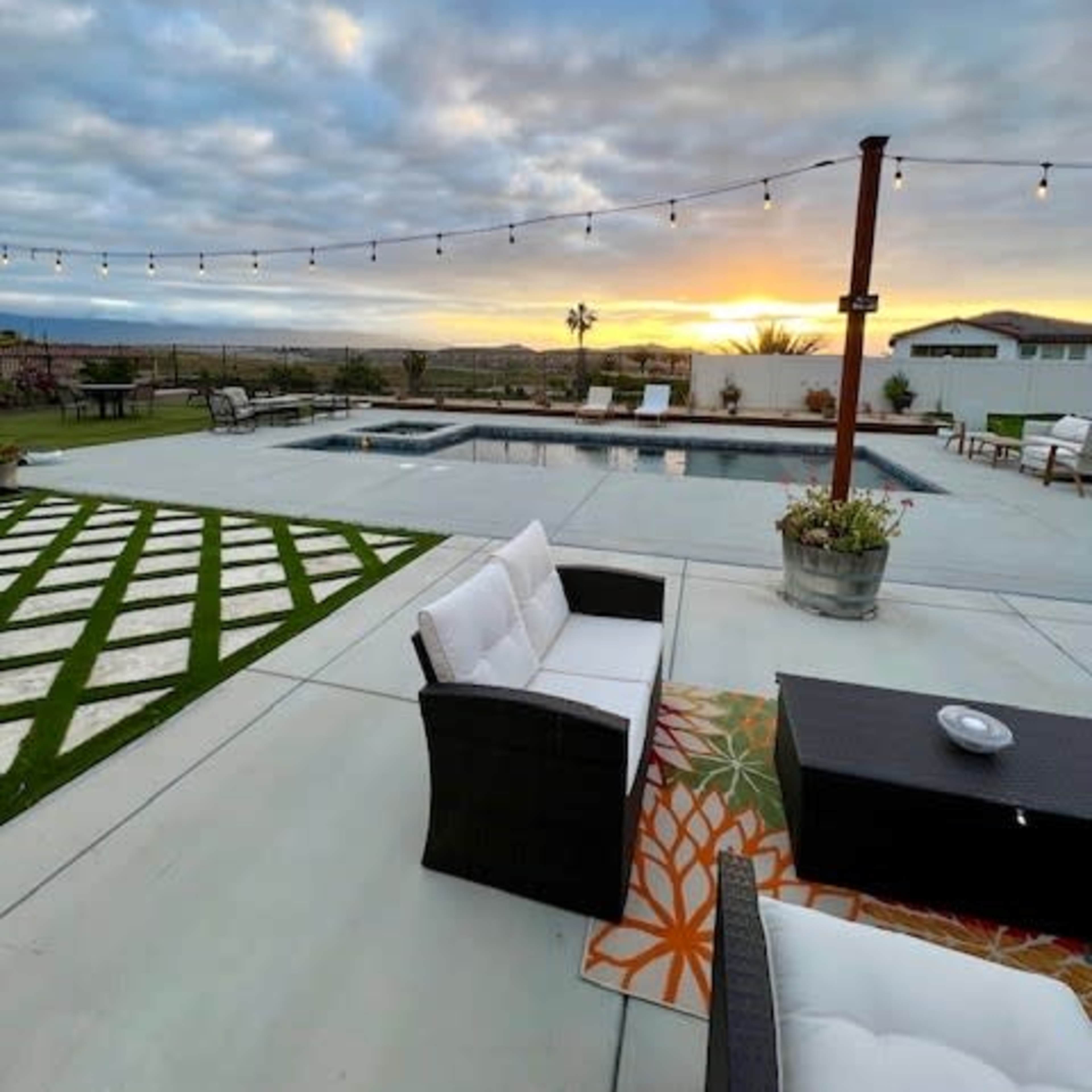 A patio area with white outdoor furniture, a swimming pool, and a sunset in the background, framed by string lights and a landscaped lawn.