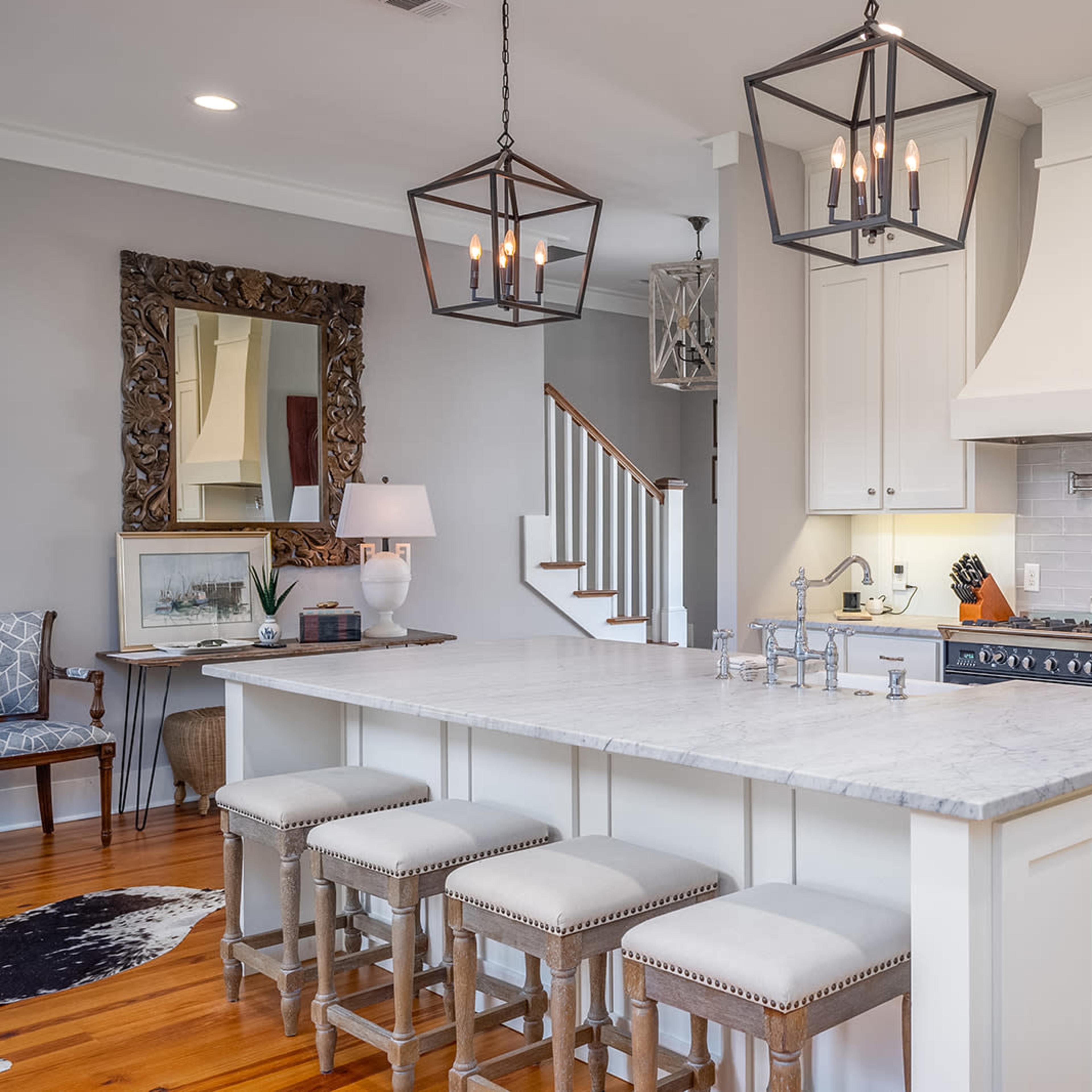 A modern kitchen features a marble island with four upholstered stools, stainless steel appliances, and white cabinetry, with a staircase and a decorative mirror in the background.