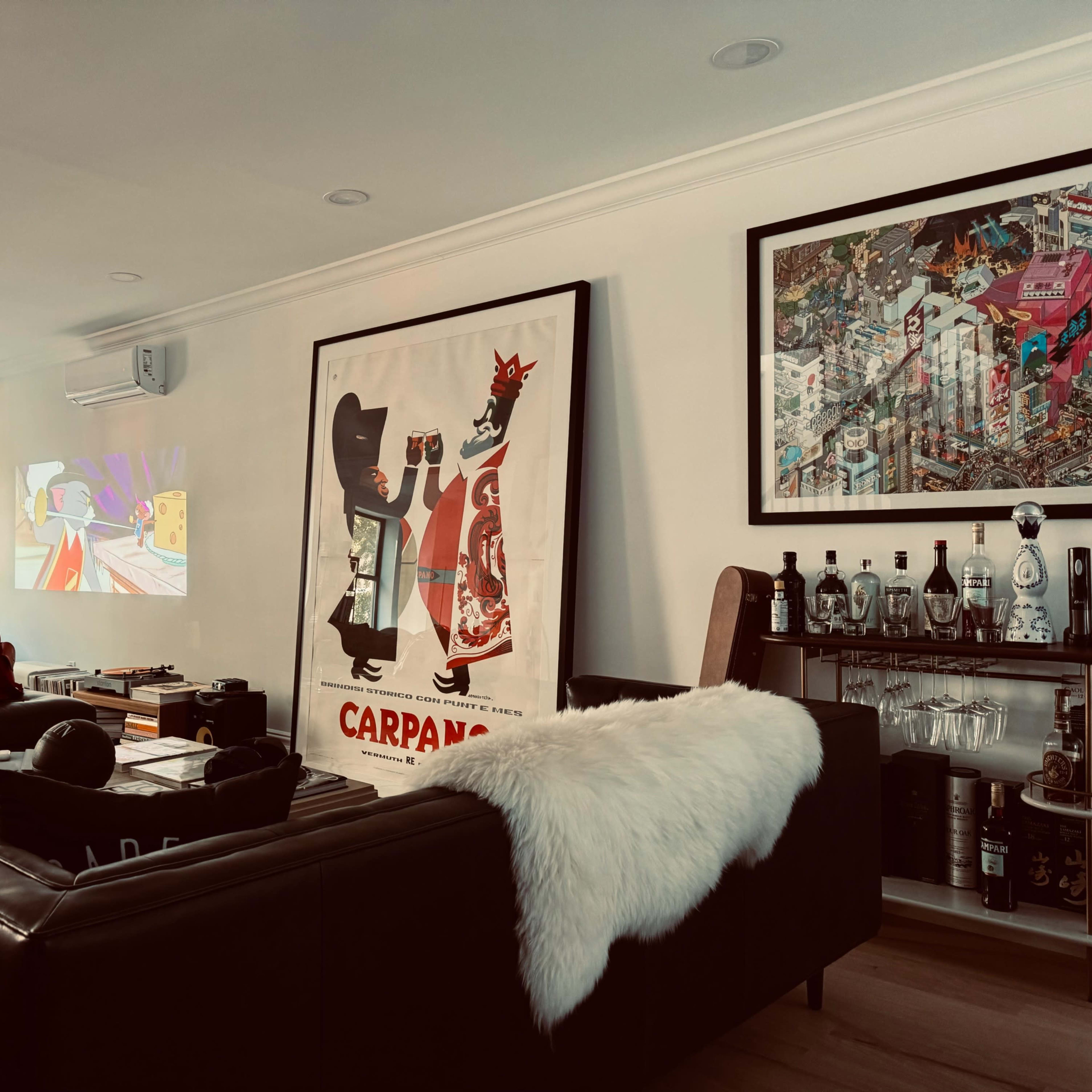 A modern living room featuring a black leather couch, various art pieces on the walls, and several items like luggage and clothing scattered around.