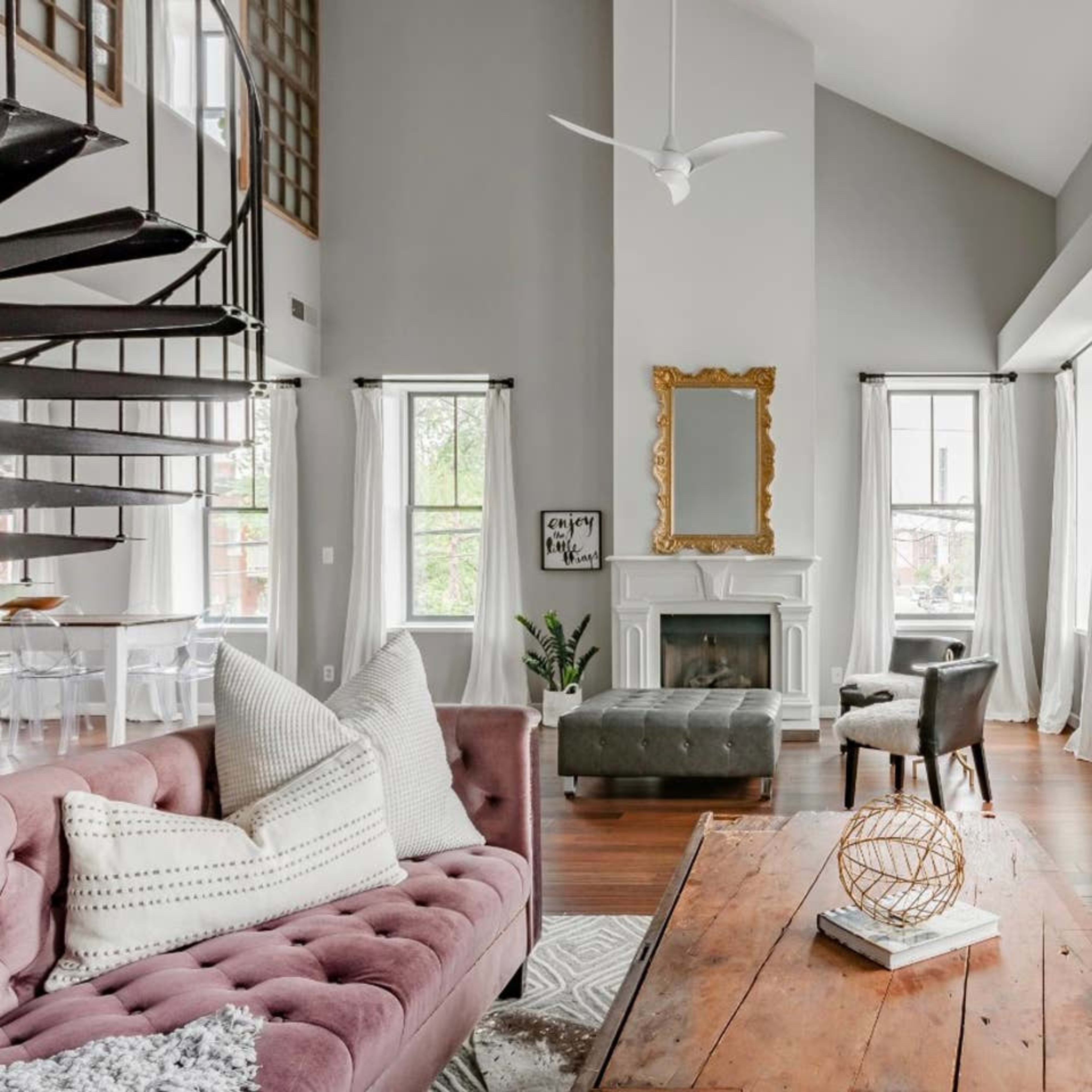 A modern living room featuring two tufted sofas, a wooden coffee table, and large windows with sheer curtains, complemented by a spiral staircase and a decorative fireplace.