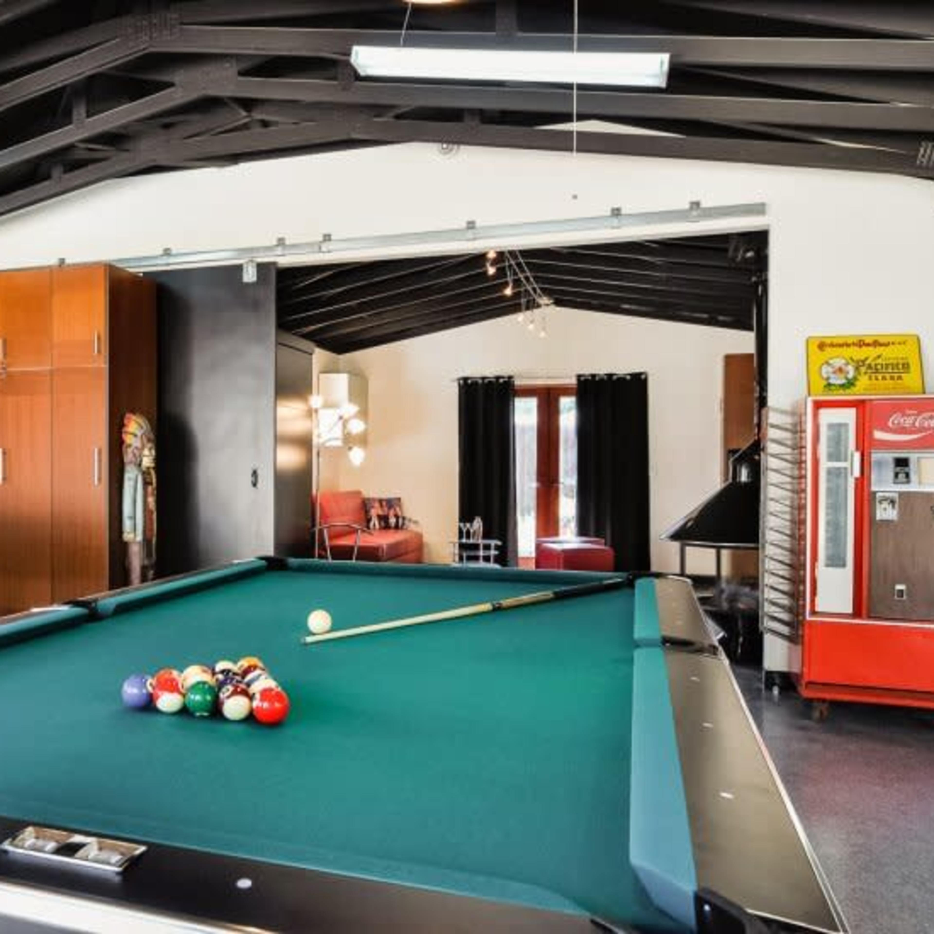 A pool table with a full rack of balls is positioned in a game room featuring high ceilings, a vintage vending machine, and a seating area.