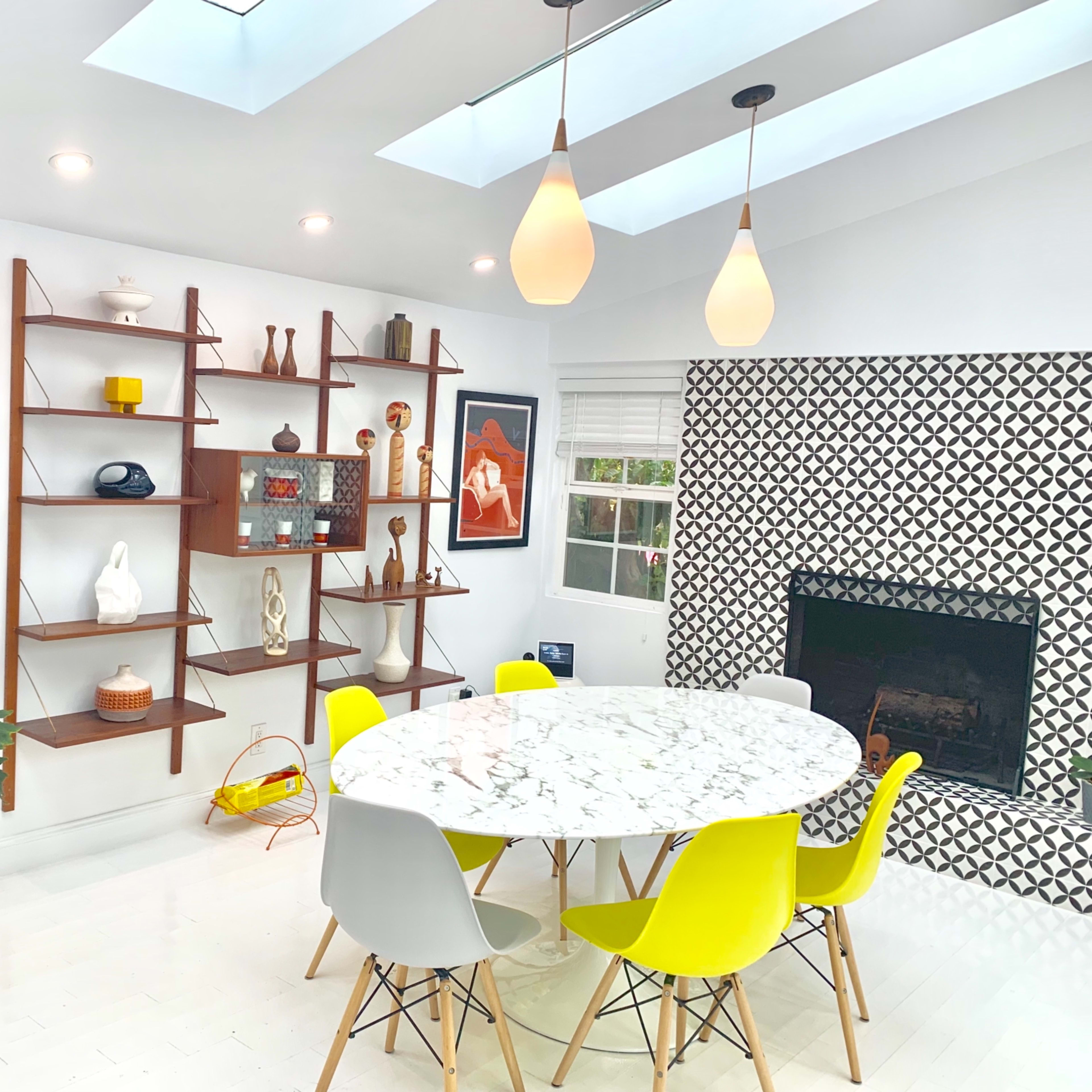 A modern dining area features a round marble table surrounded by colorful chairs, a decorative bookshelf, and a geometric patterned accent wall.