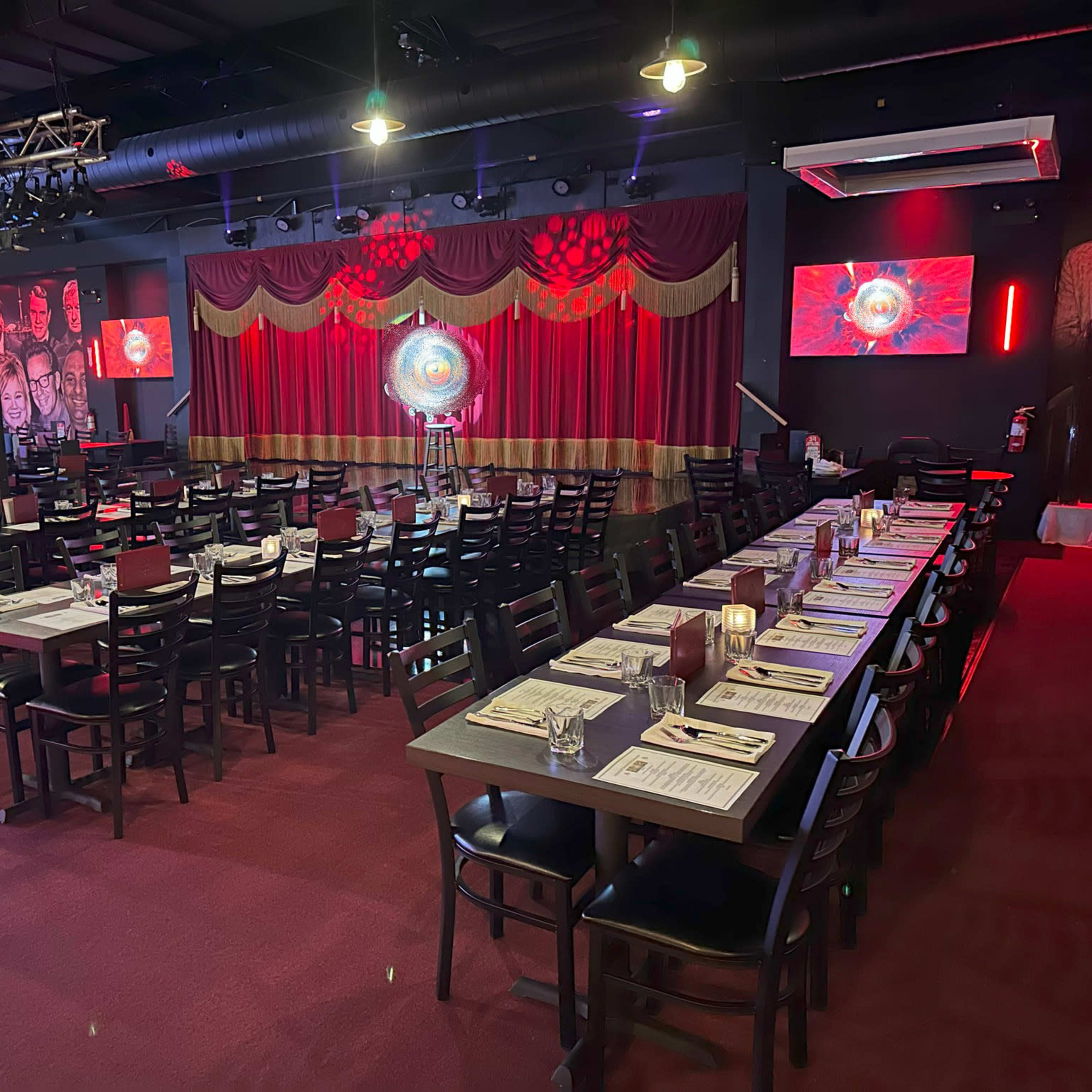 An elegantly arranged restaurant interior featuring rows of tables set for dining, red drapes on the stage, and colorful lighting.