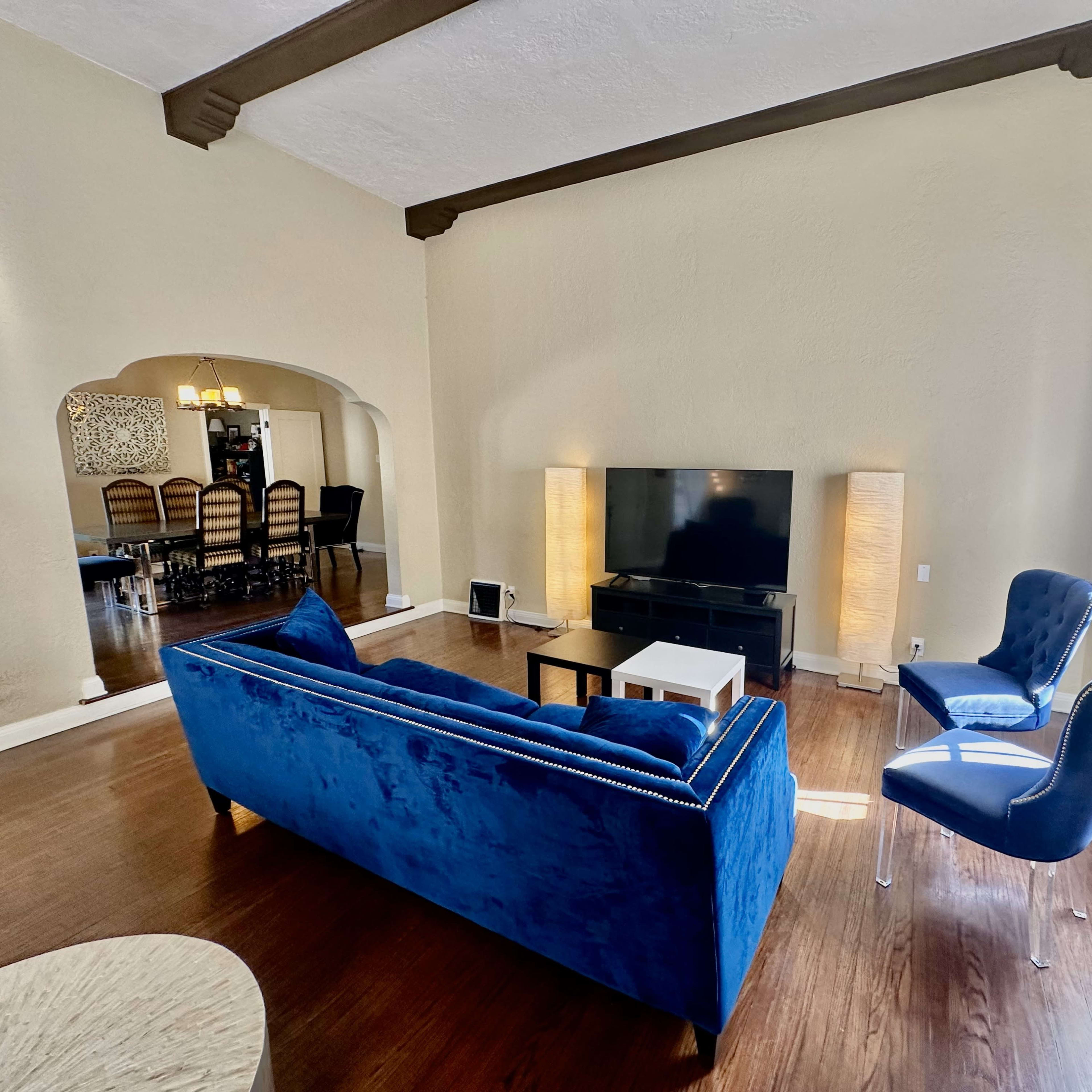 A modern living room features a blue velvet sofa, a black television stand, and two floor lamps, with a dining area visible in the background.