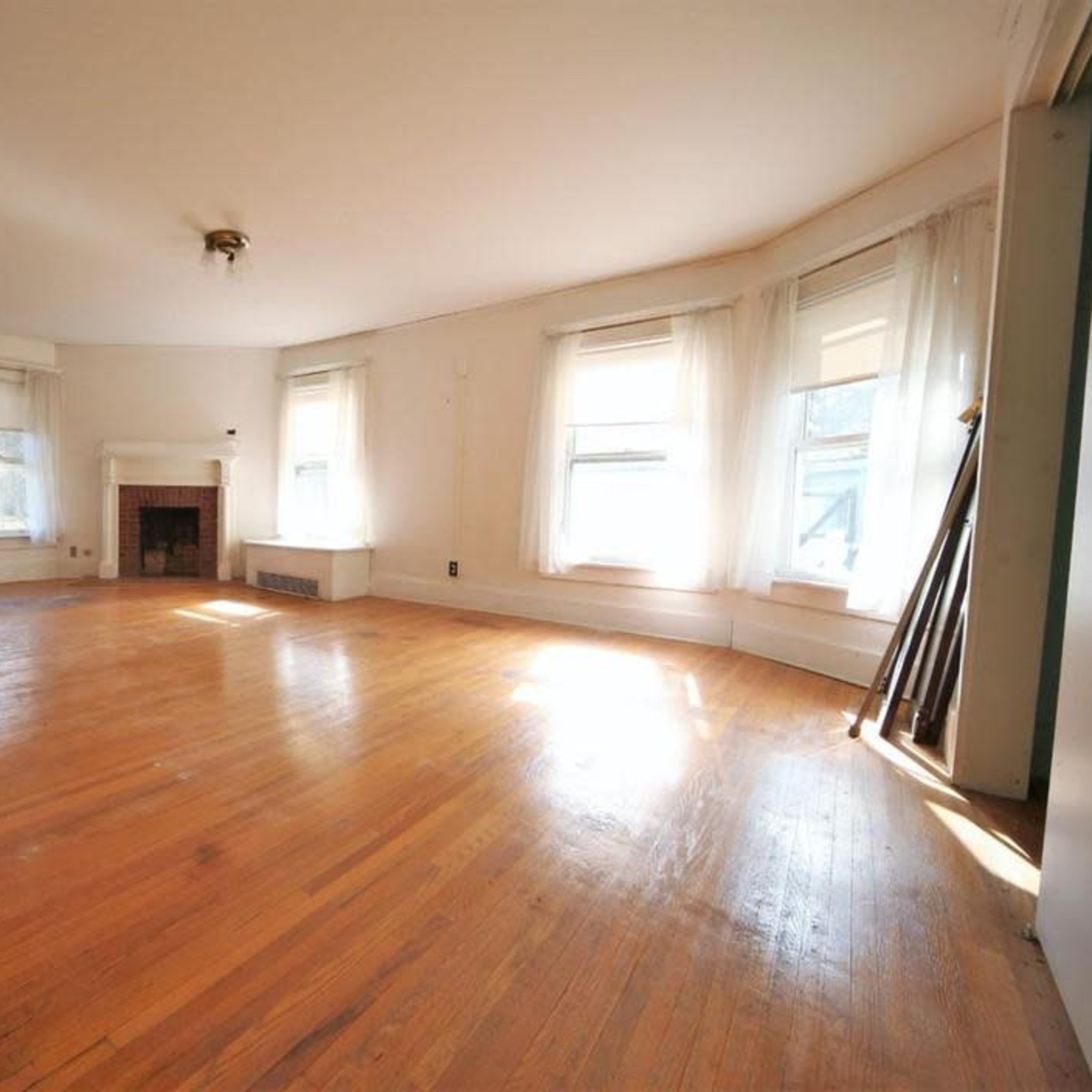 A bright, empty living room with hardwood floors, large windows, and a fireplace.