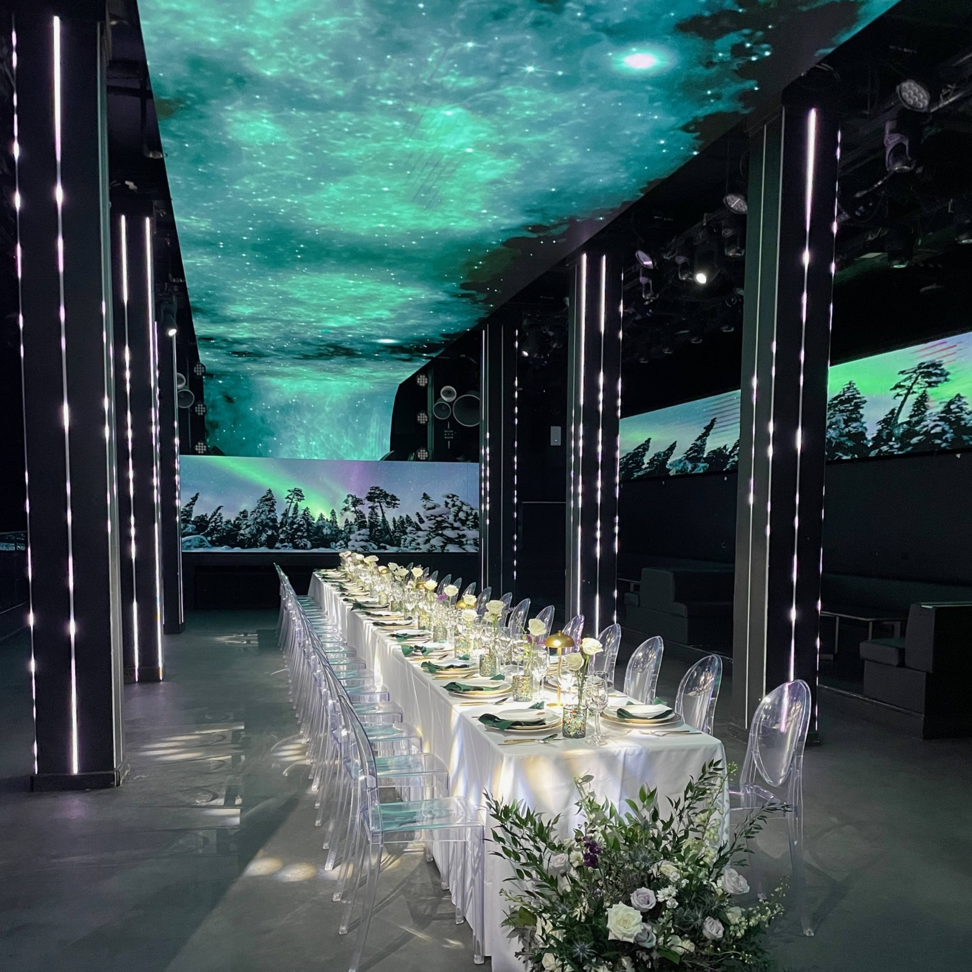 A long dining table is set with plates and glasses under a ceiling projection of a starry night sky, surrounded by greenery and modern decor.