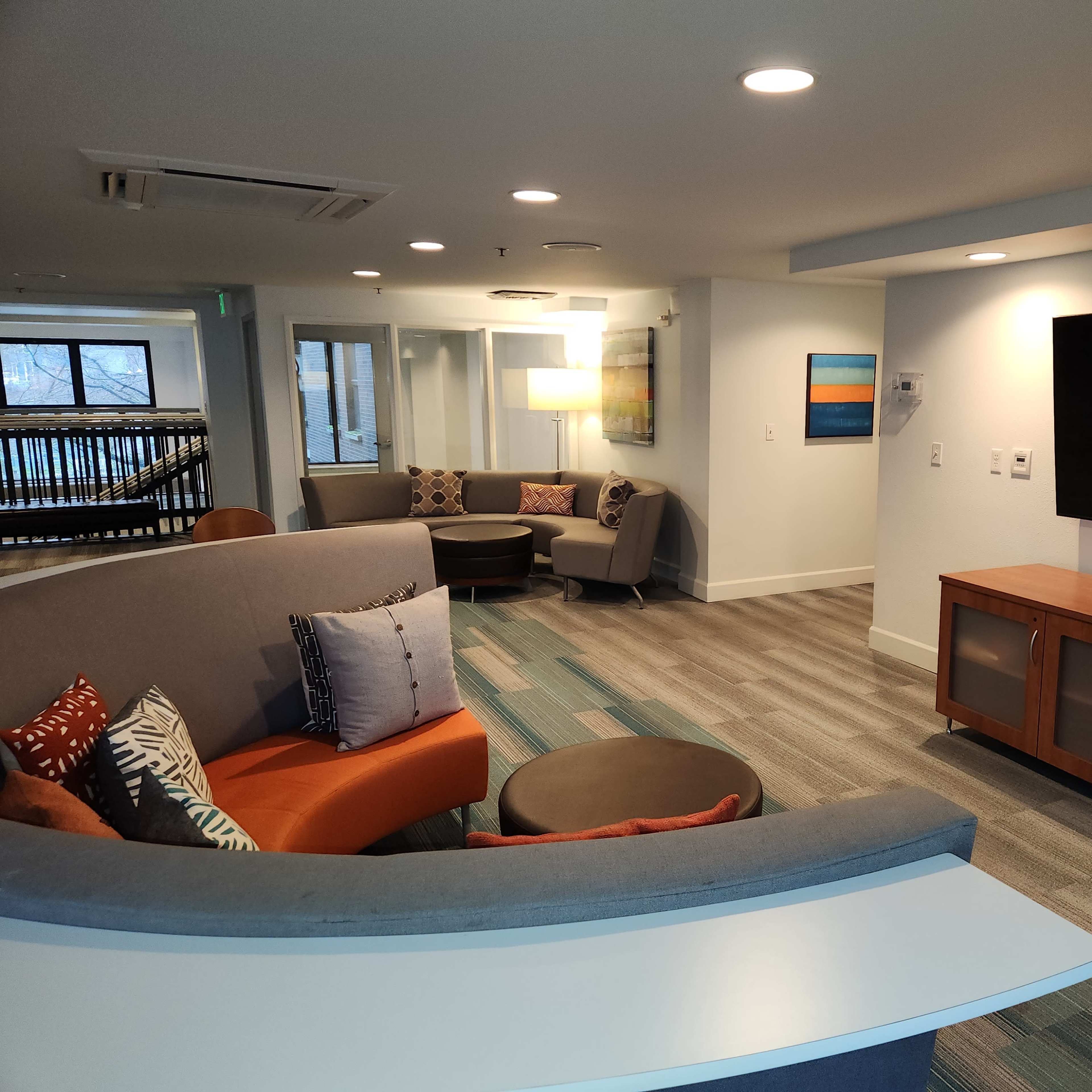 A modern lounge area featuring a curved gray couch, a wooden television cabinet, and colorful cushions, with a flat-screen TV mounted on the wall.