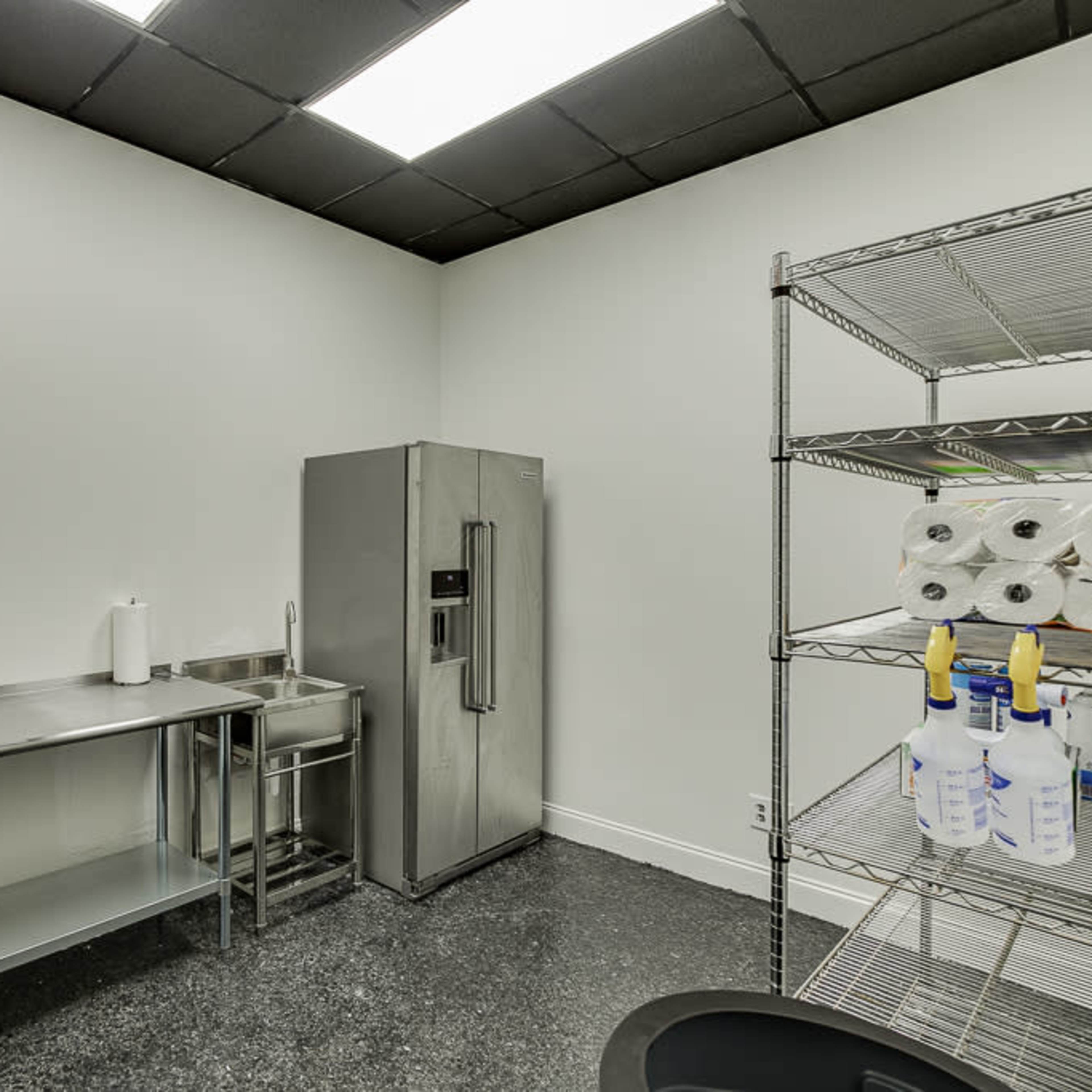A clean, empty break room with a stainless steel table, a refrigerator, and a storage shelf with cleaning supplies.