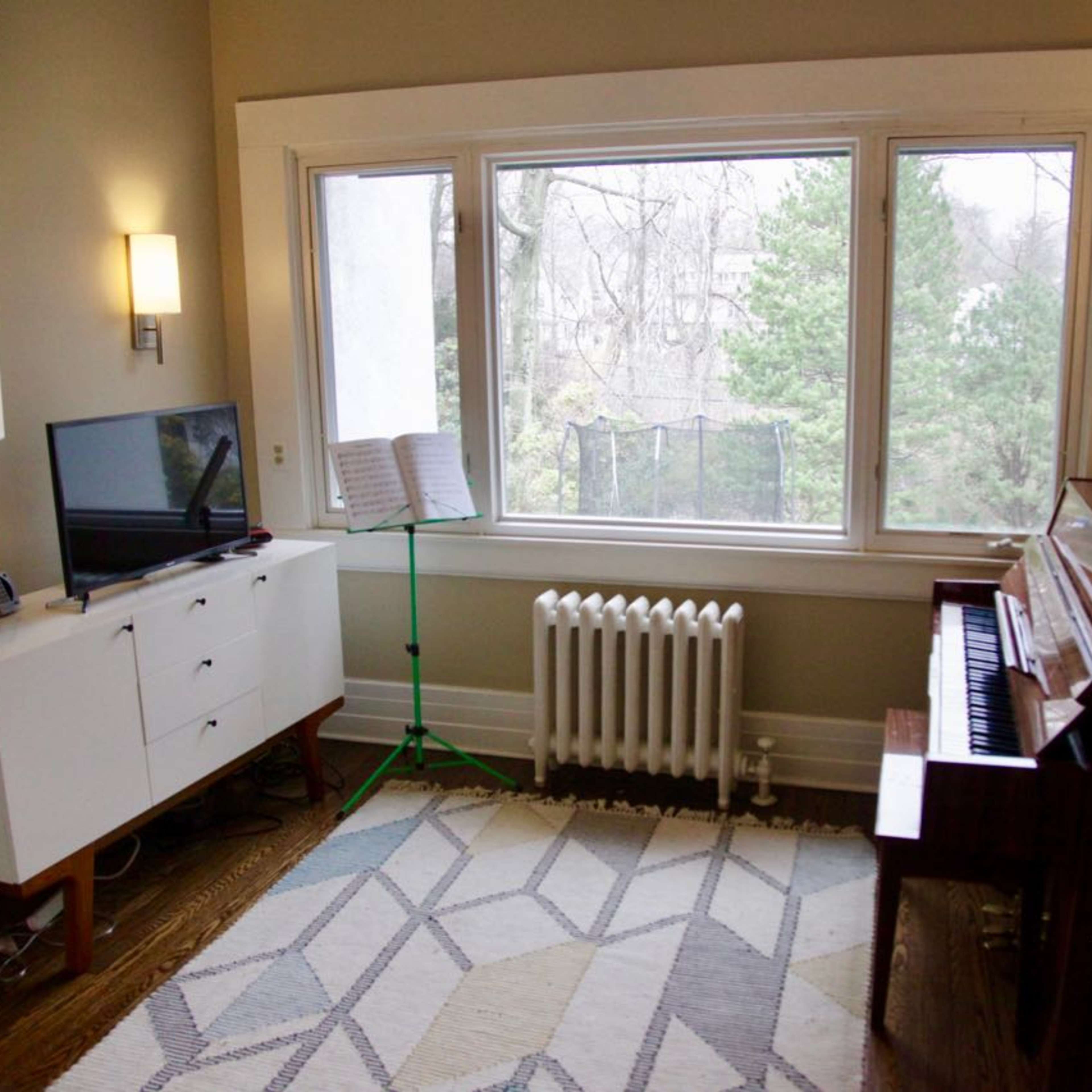 A room features a piano, a TV on a white cabinet, a music stand, and a large window with a view of trees outside.
