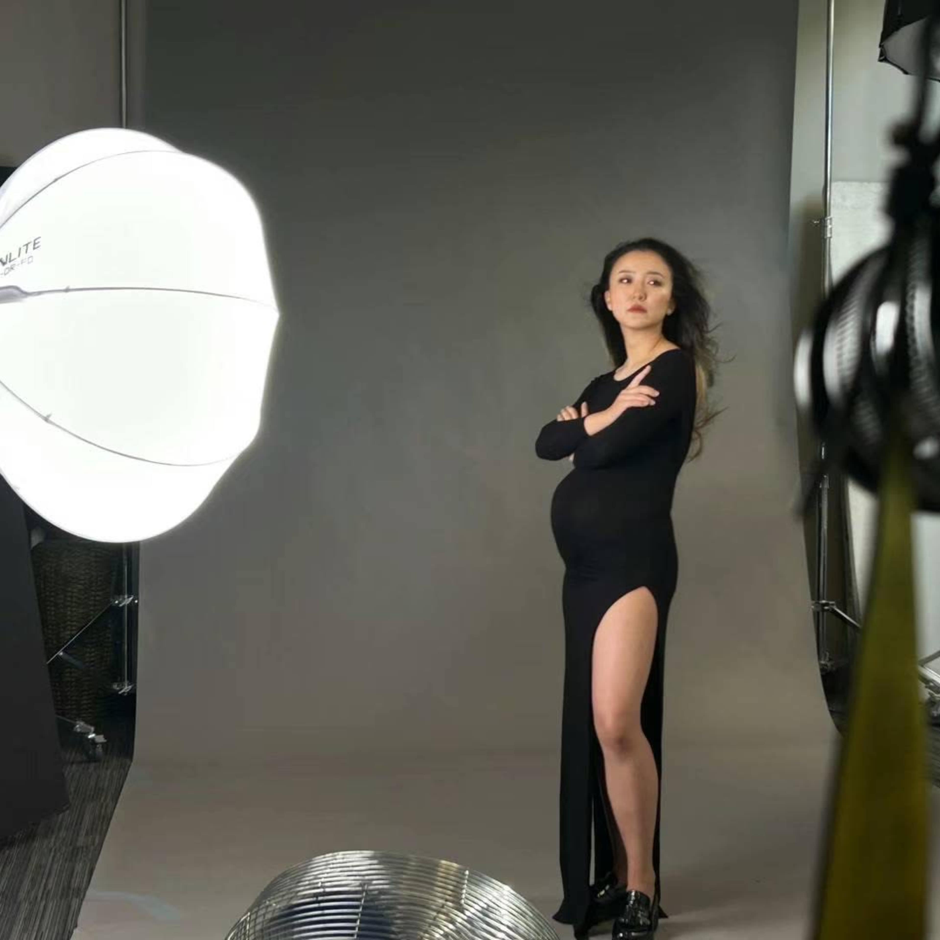 A pregnant woman in a black dress poses confidently with her arms crossed in a photography studio.