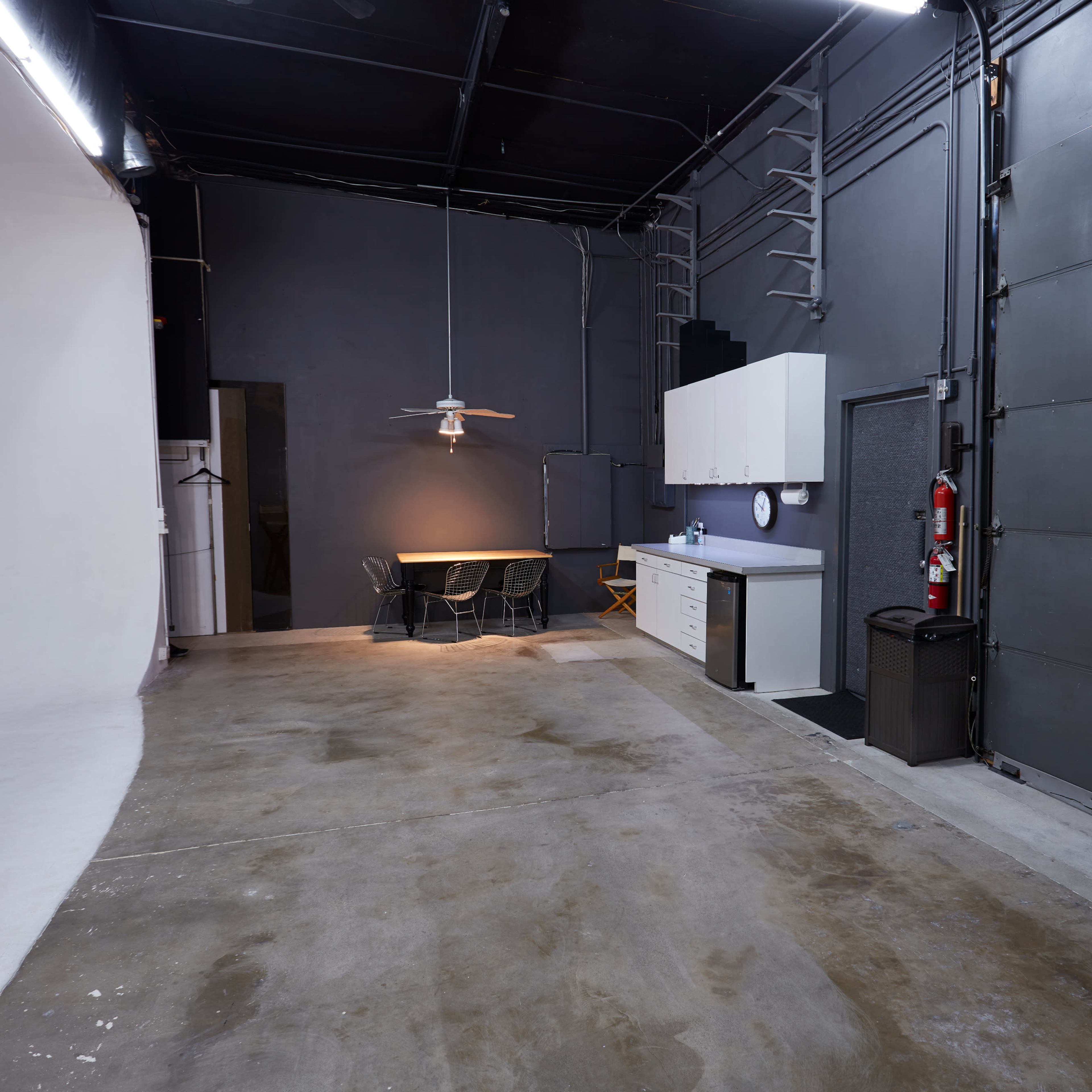 A spacious studio with a white backdrop on one side, a table and chairs, and a kitchenette area against a gray wall.