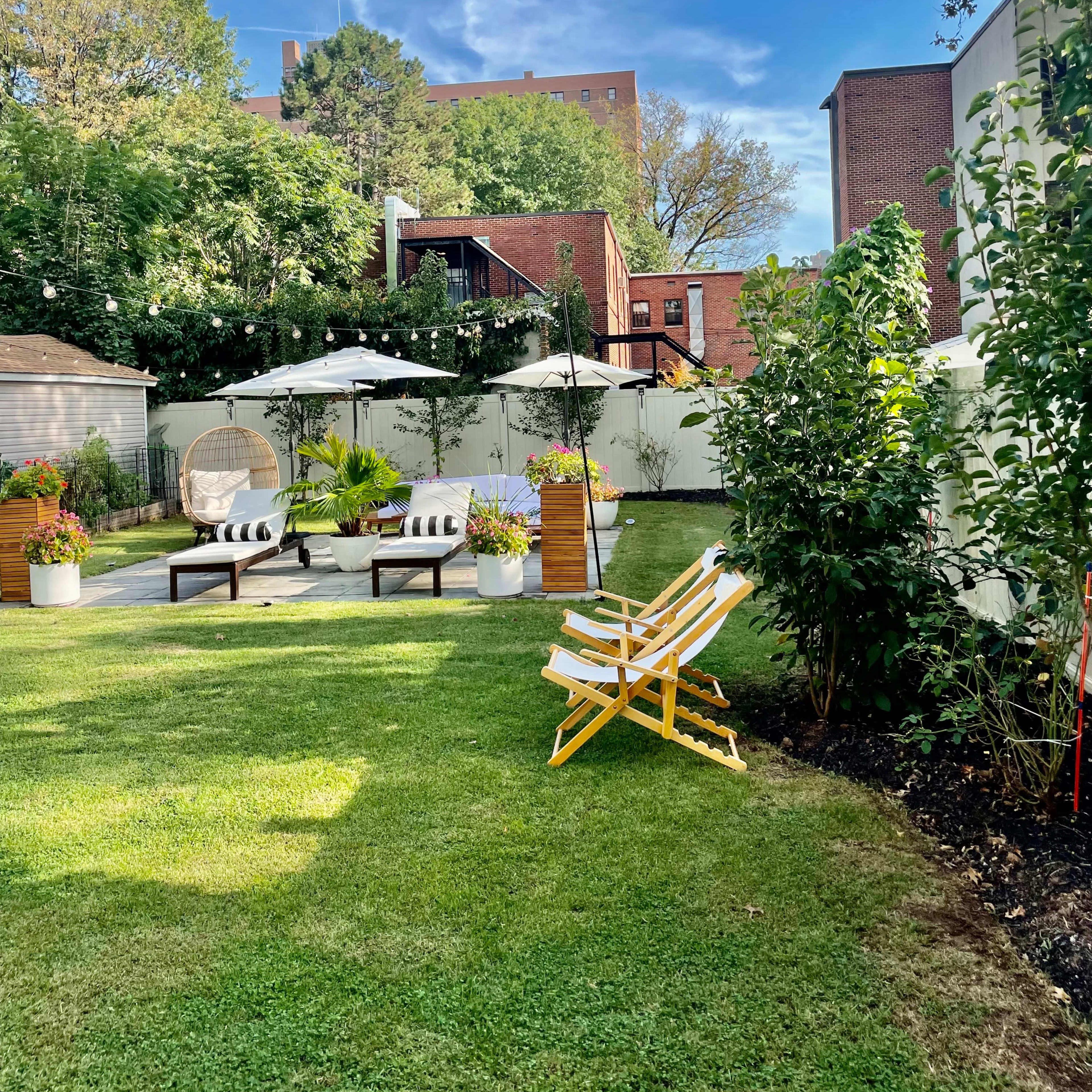 A landscaped backyard with seating areas, umbrellas, and string lights set against a blue sky and greenery.