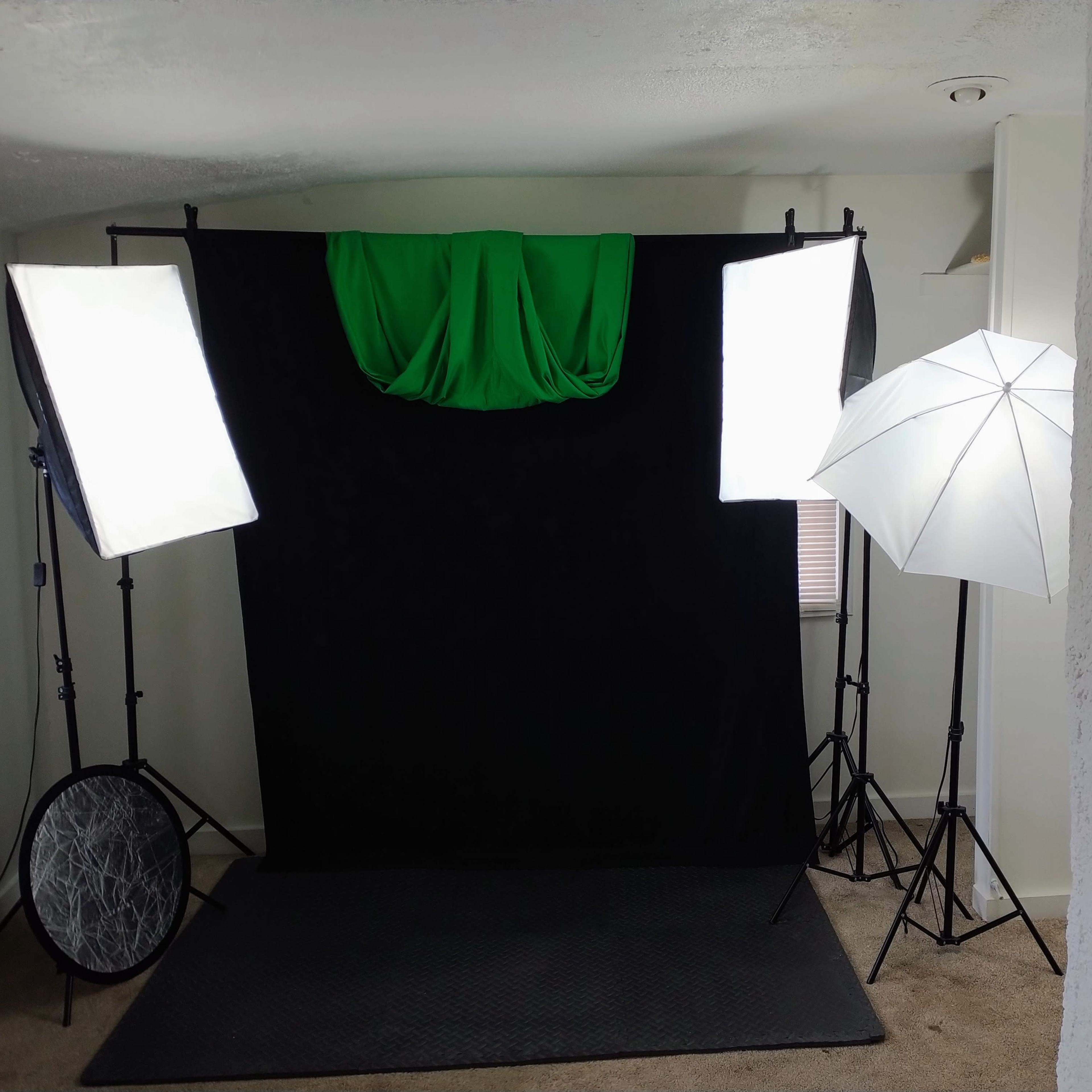 A photo studio setup featuring a black backdrop, green drapery, and three lighting sources, including two softboxes and one umbrella light.