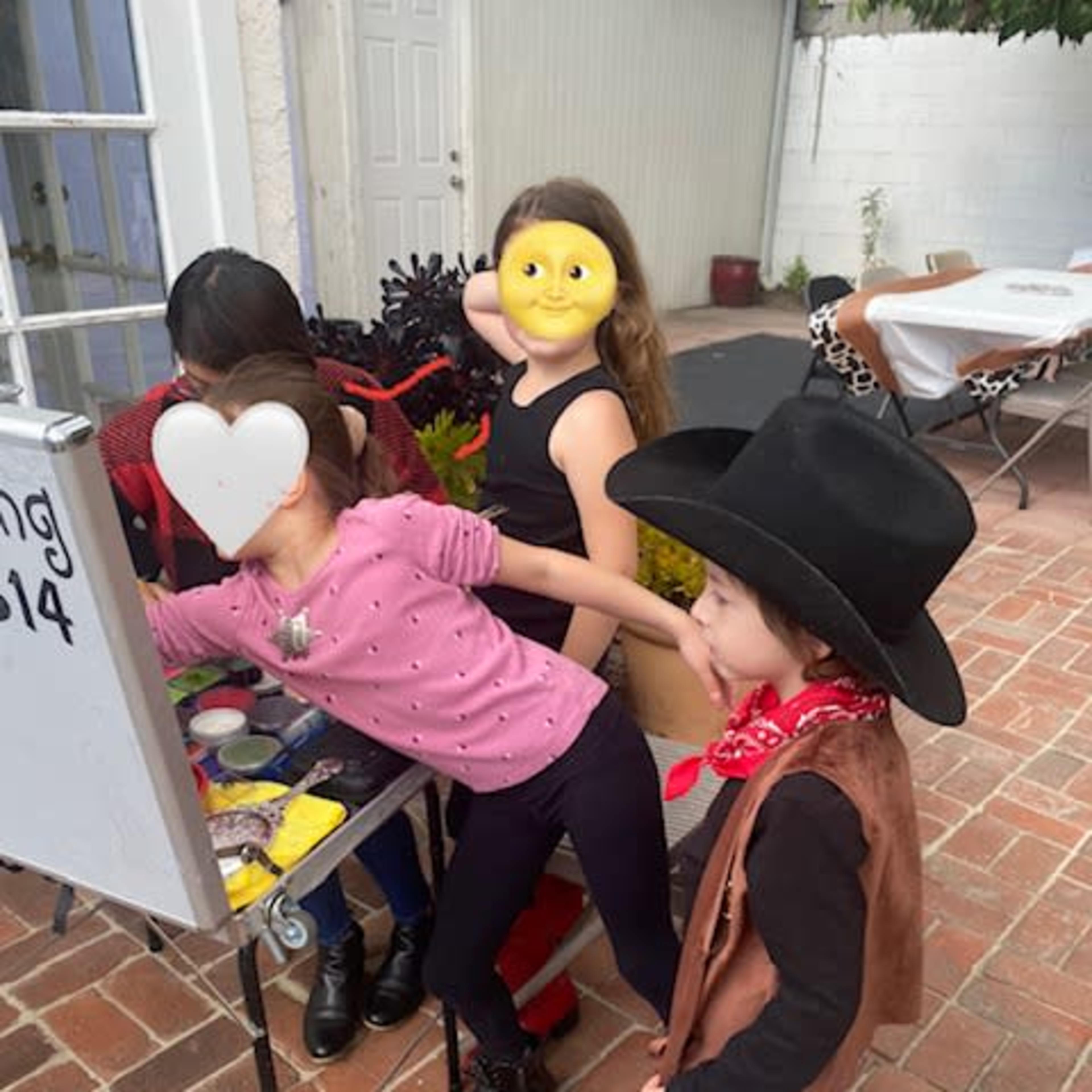 A group of children engages in a craft activity outdoors, with one child wearing a yellow mask and another in a cowboy costume.