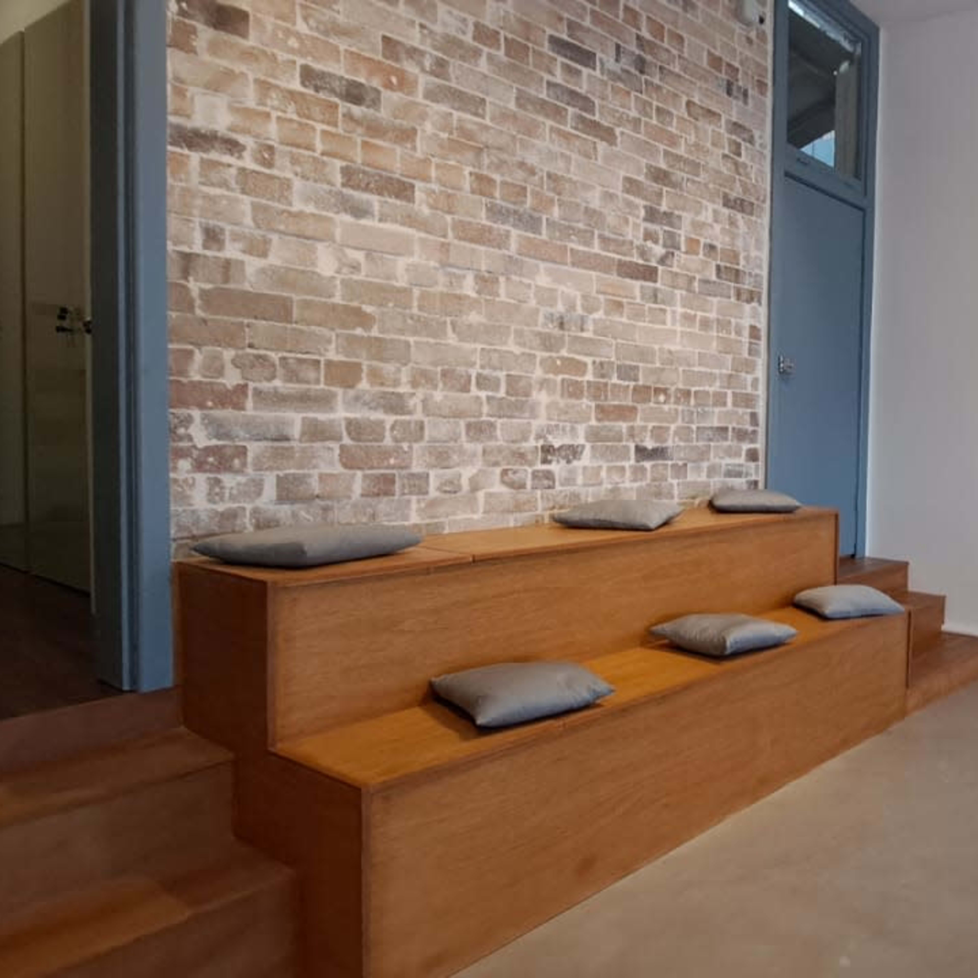 A minimalistic interior space featuring a wooden bench with cushions against a brick wall and concrete flooring.