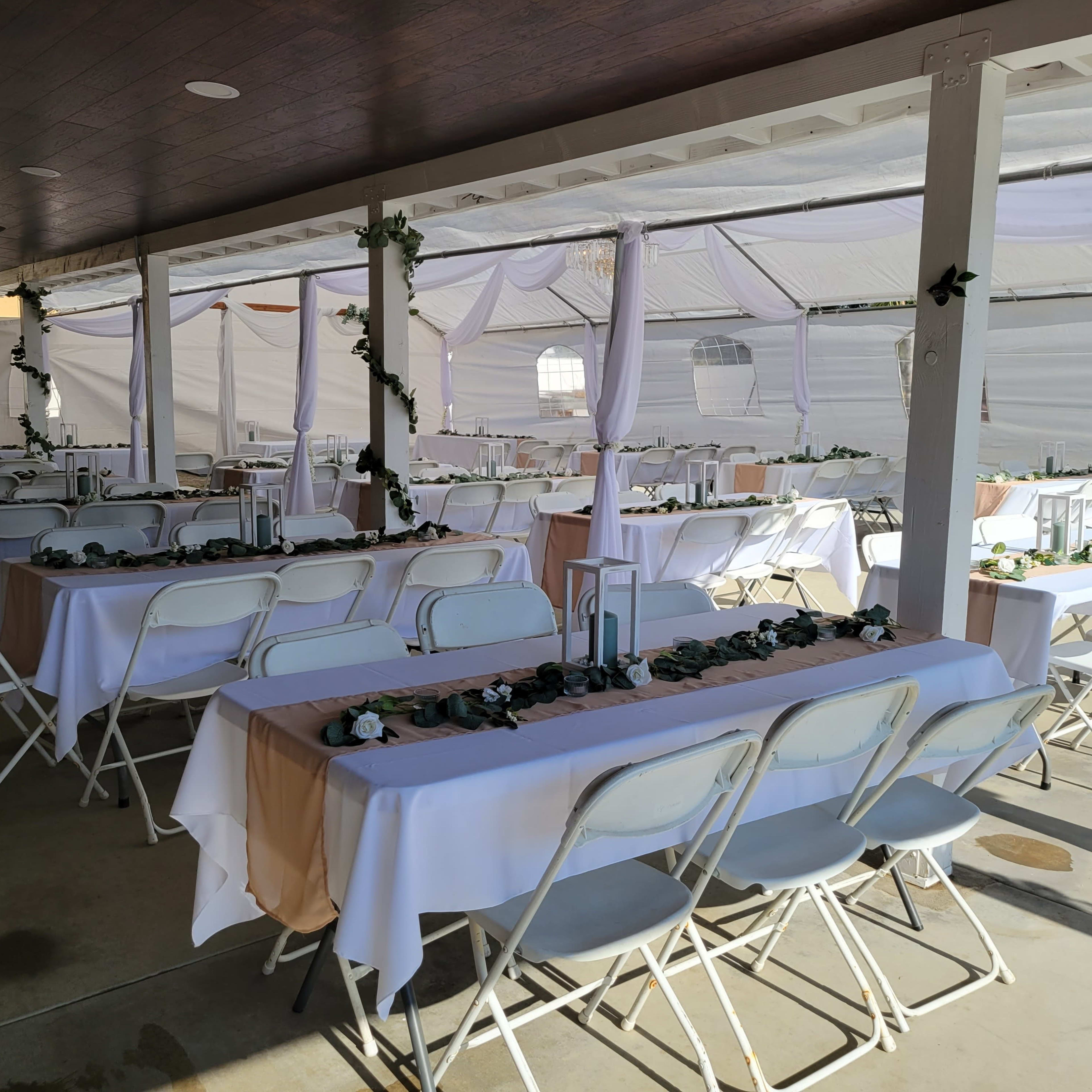 A covered outdoor venue with several rows of tables set with white tablecloths and decorated with greenery.