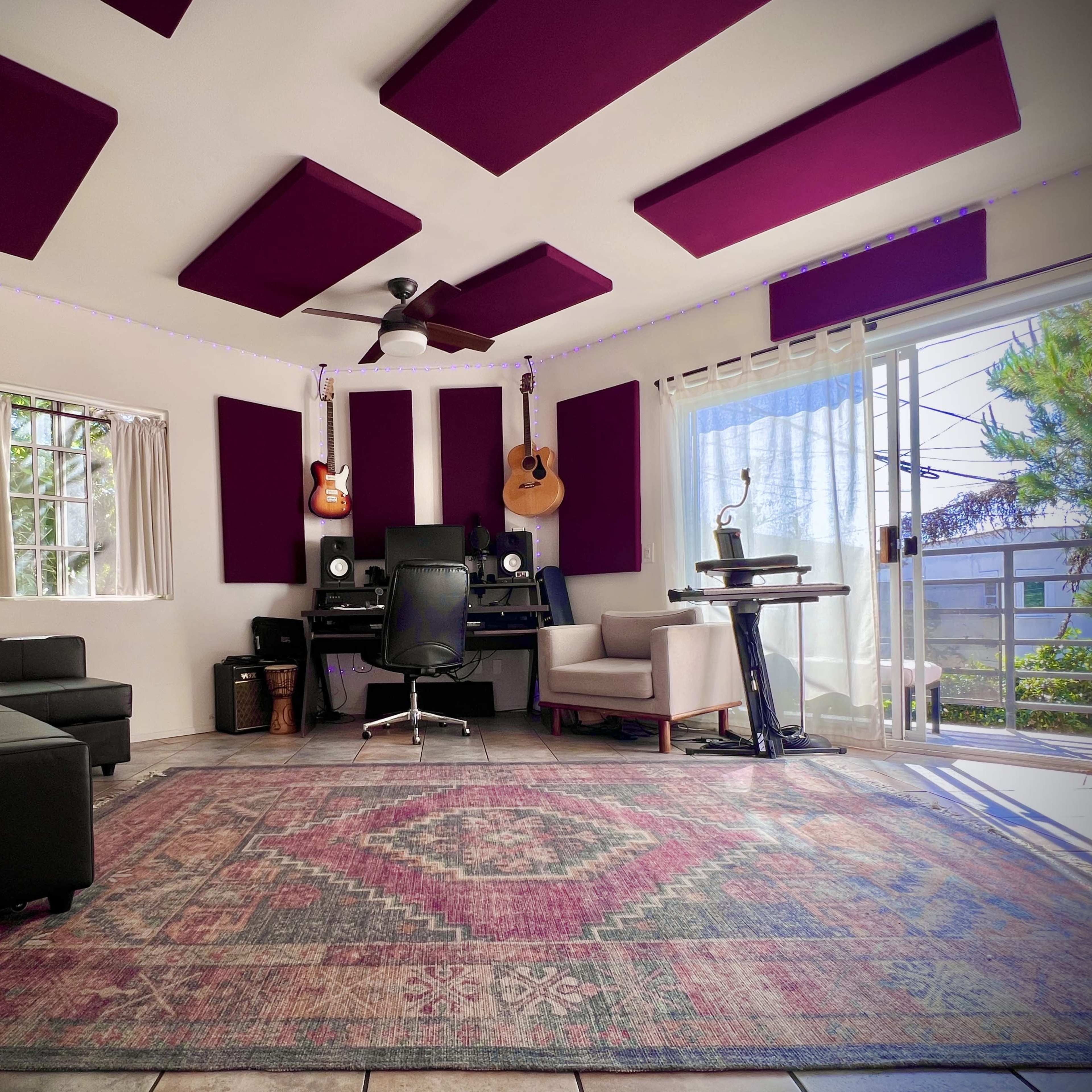 A home studio with purple acoustic panels, a desk setup with a computer, guitars on the wall, and a cozy seating area.