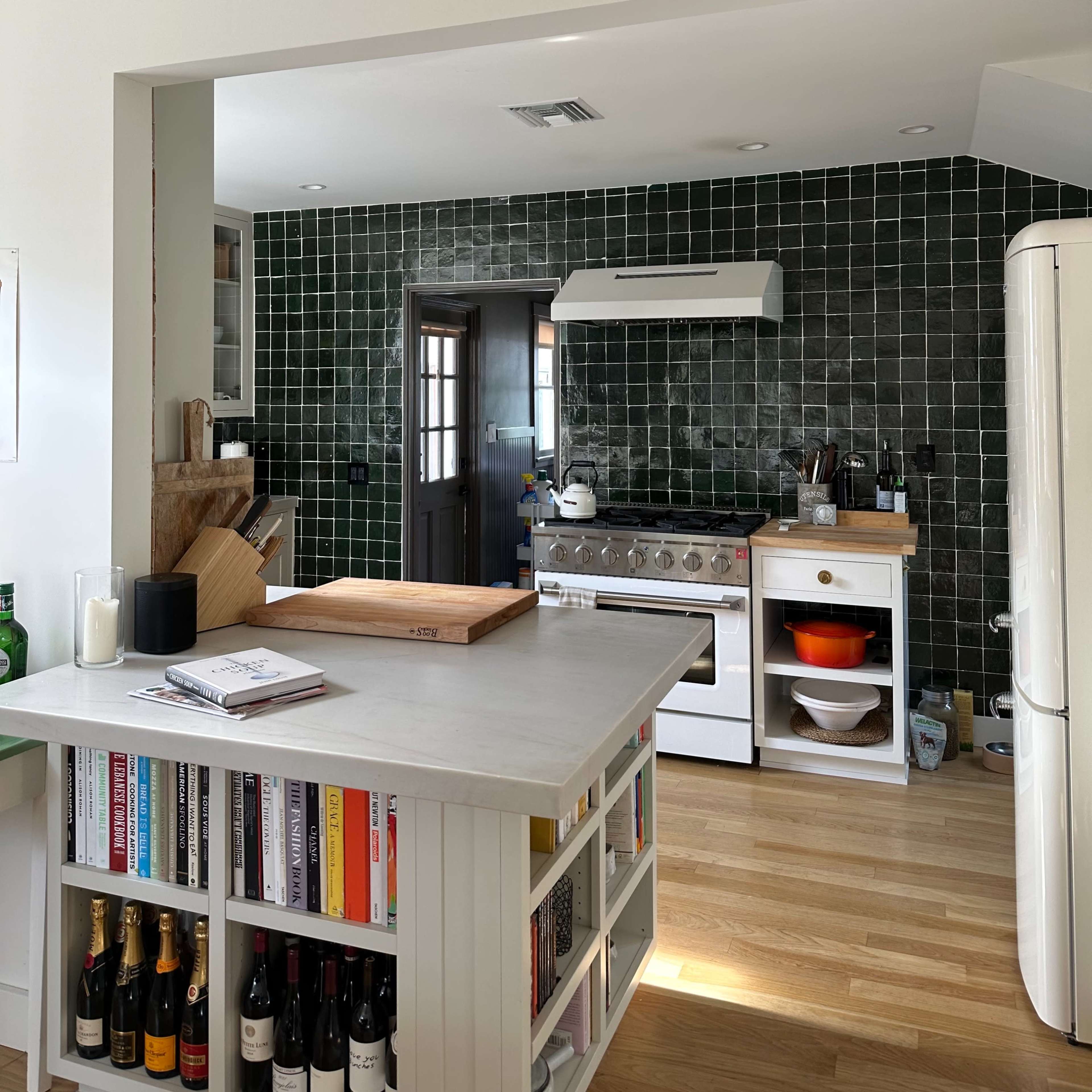 A modern kitchen featuring a green tile backsplash, a stainless steel stove, and a white refrigerator, with a wooden countertop holding a cutting board.