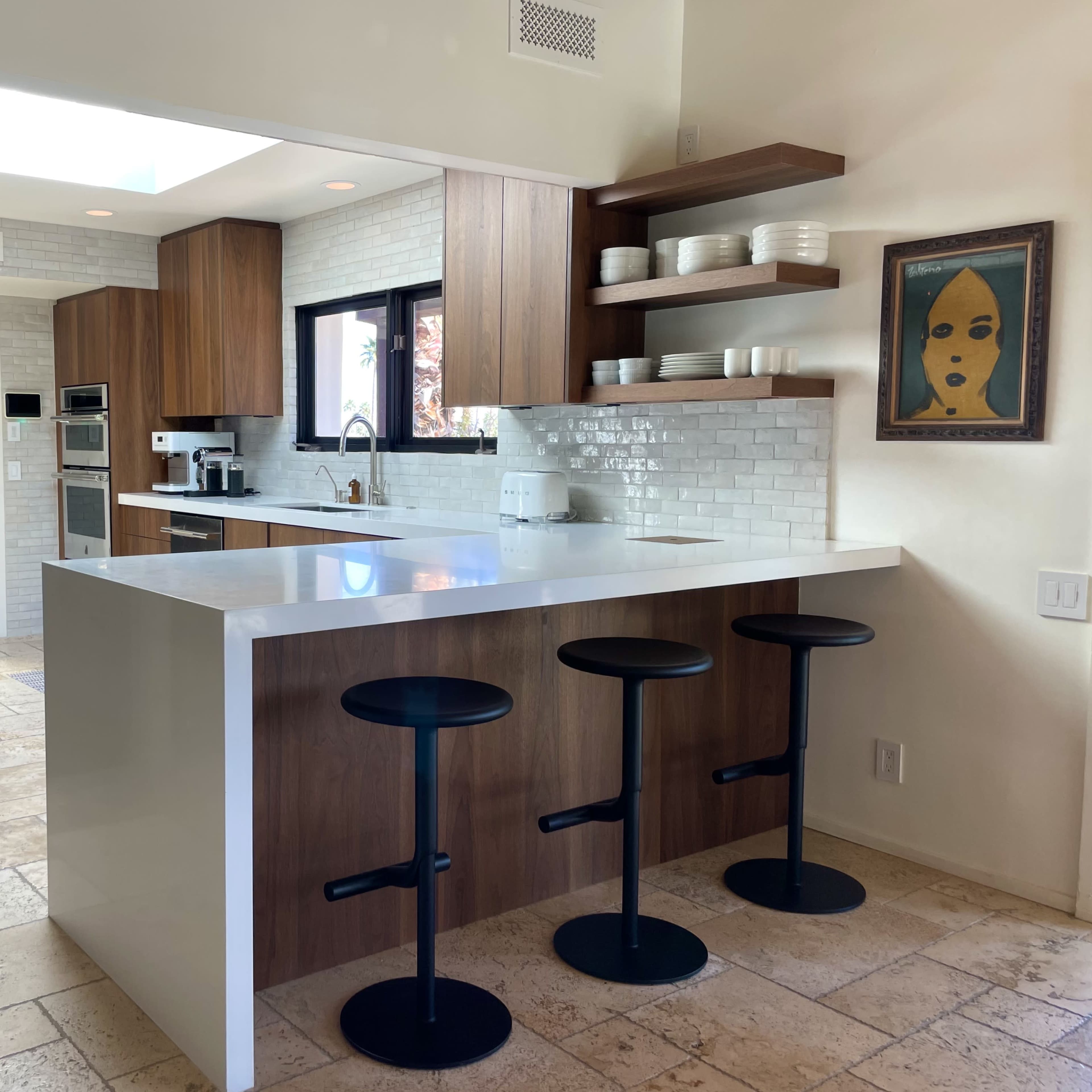 A modern kitchen featuring a white countertop island with three black stools, wooden cabinetry, and a tiled backsplash.