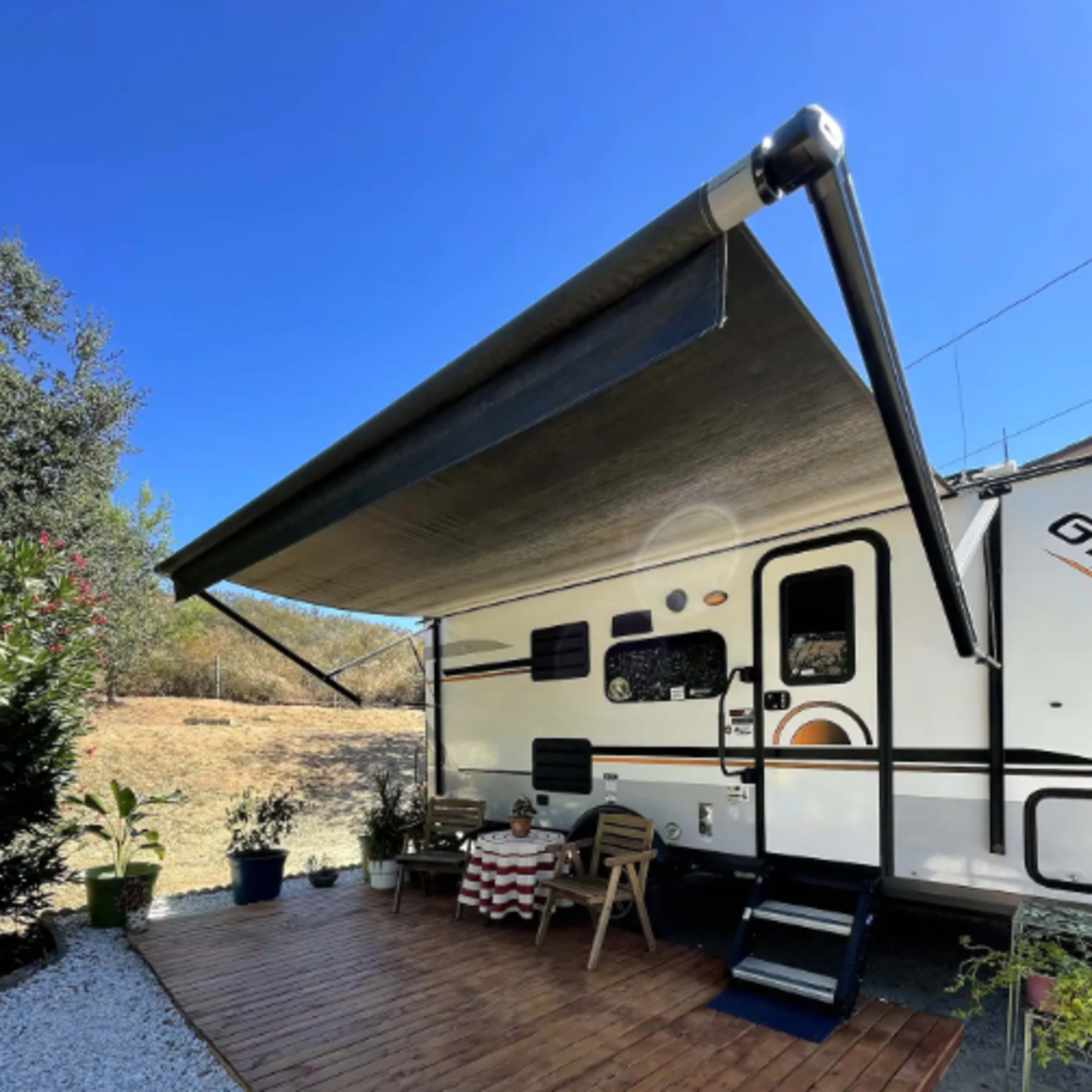 A recreational vehicle with an extended awning sits on a wooden deck surrounded by potted plants and gravel.