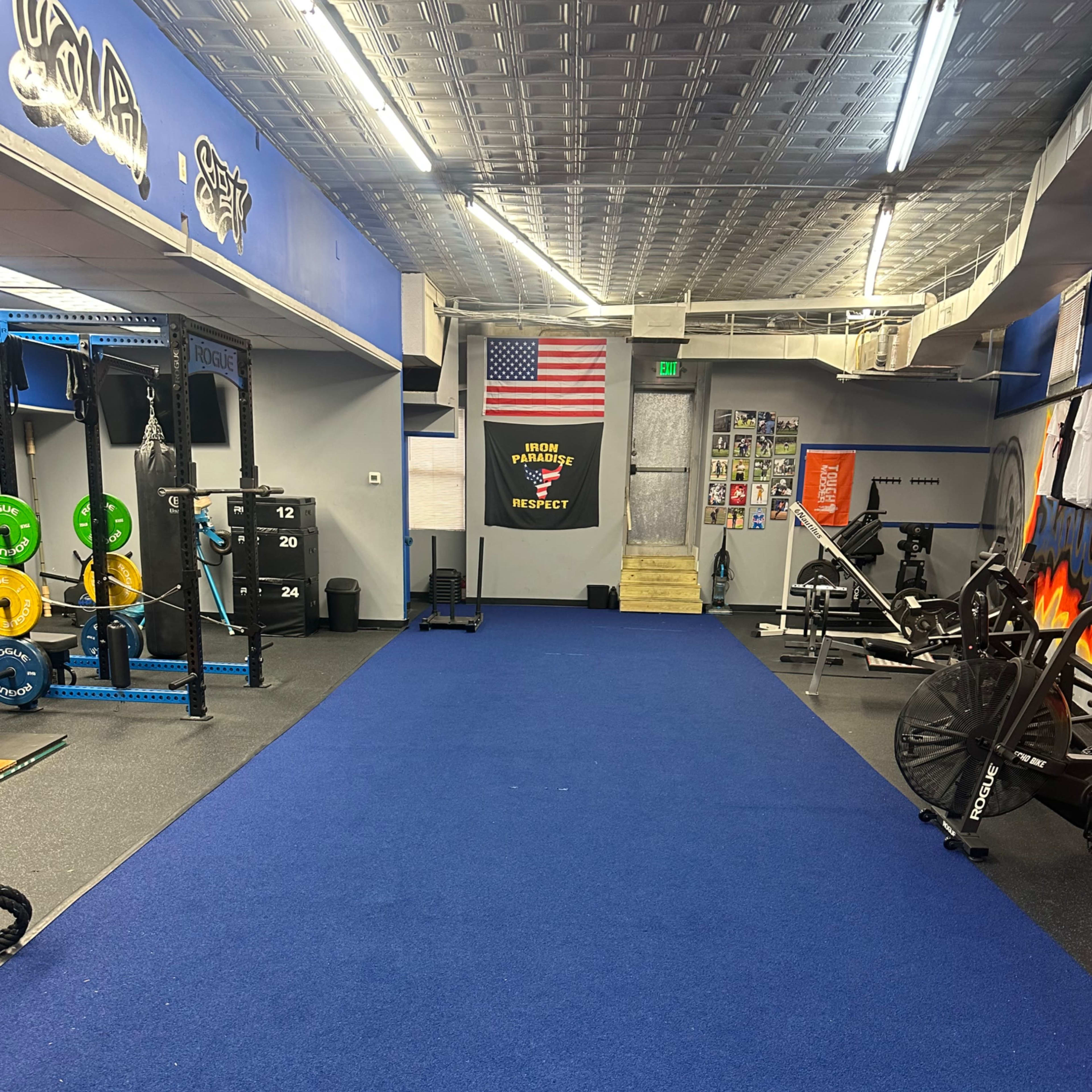 A gym interior with blue flooring, weightlifting equipment on both sides, and an American flag displayed on the wall.