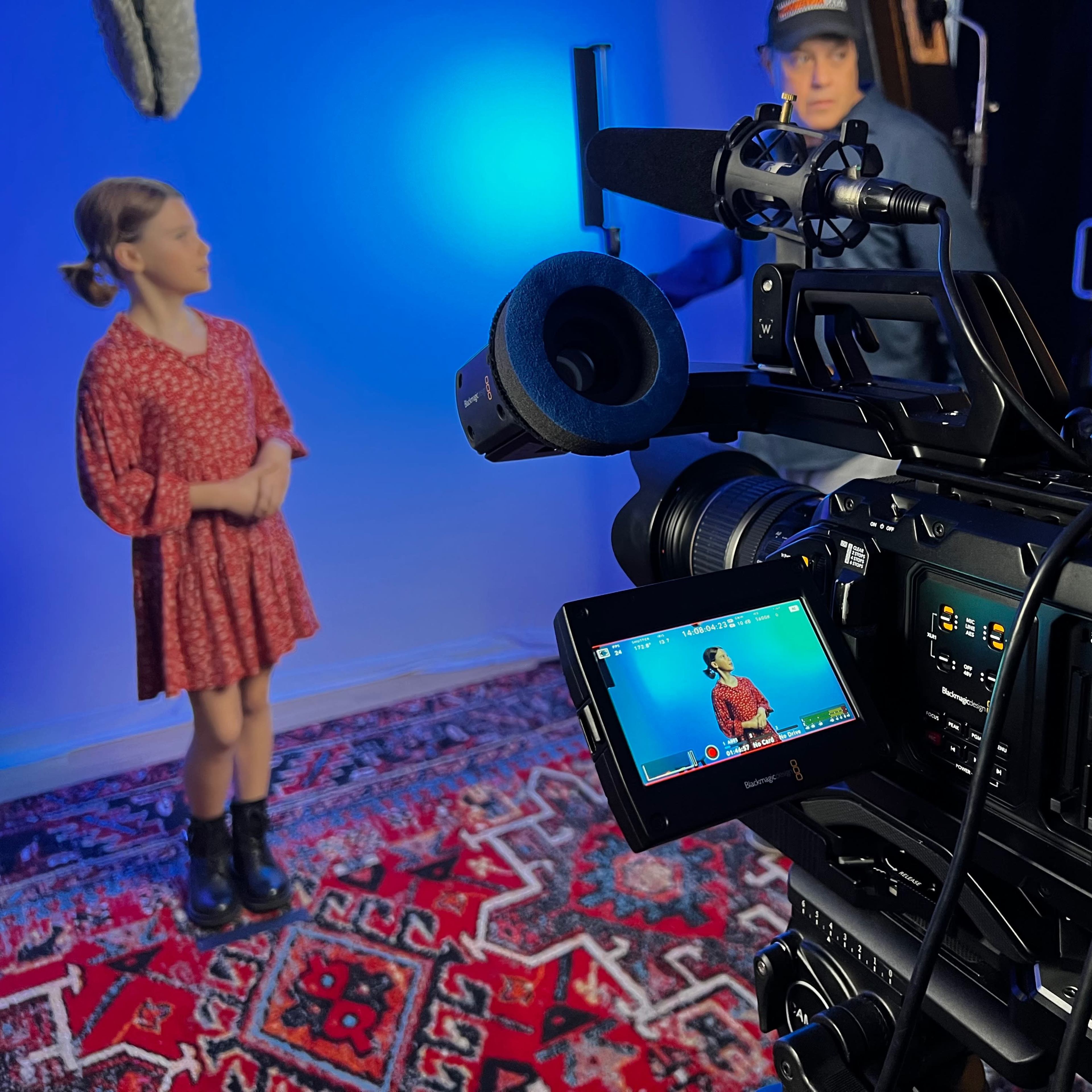 A young girl in a red dress stands in front of a blue backdrop while a camera crew prepares for a shoot.