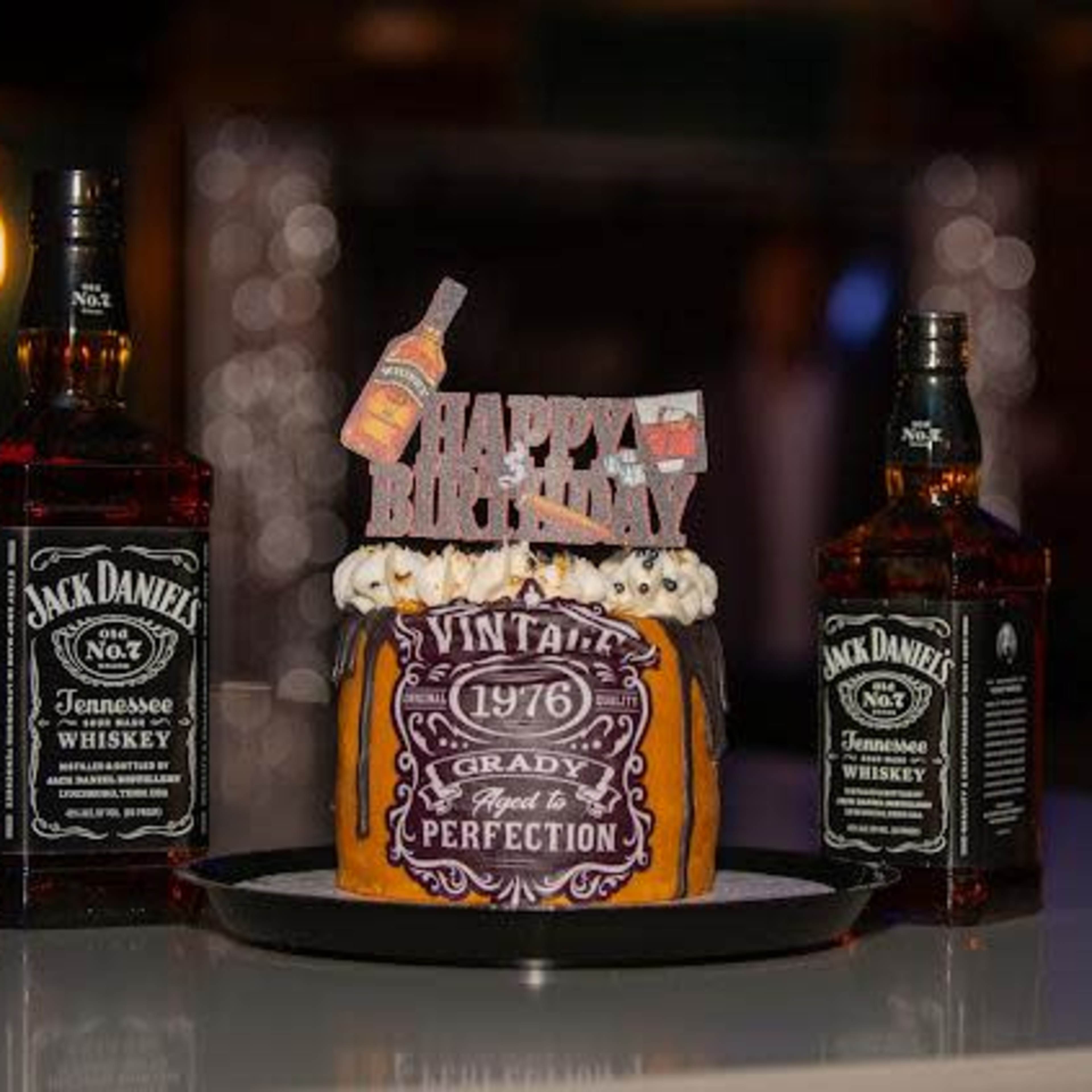 A birthday cake decorated with a "Happy Birthday" topper is flanked by two bottles of Jack Daniel's whiskey.
