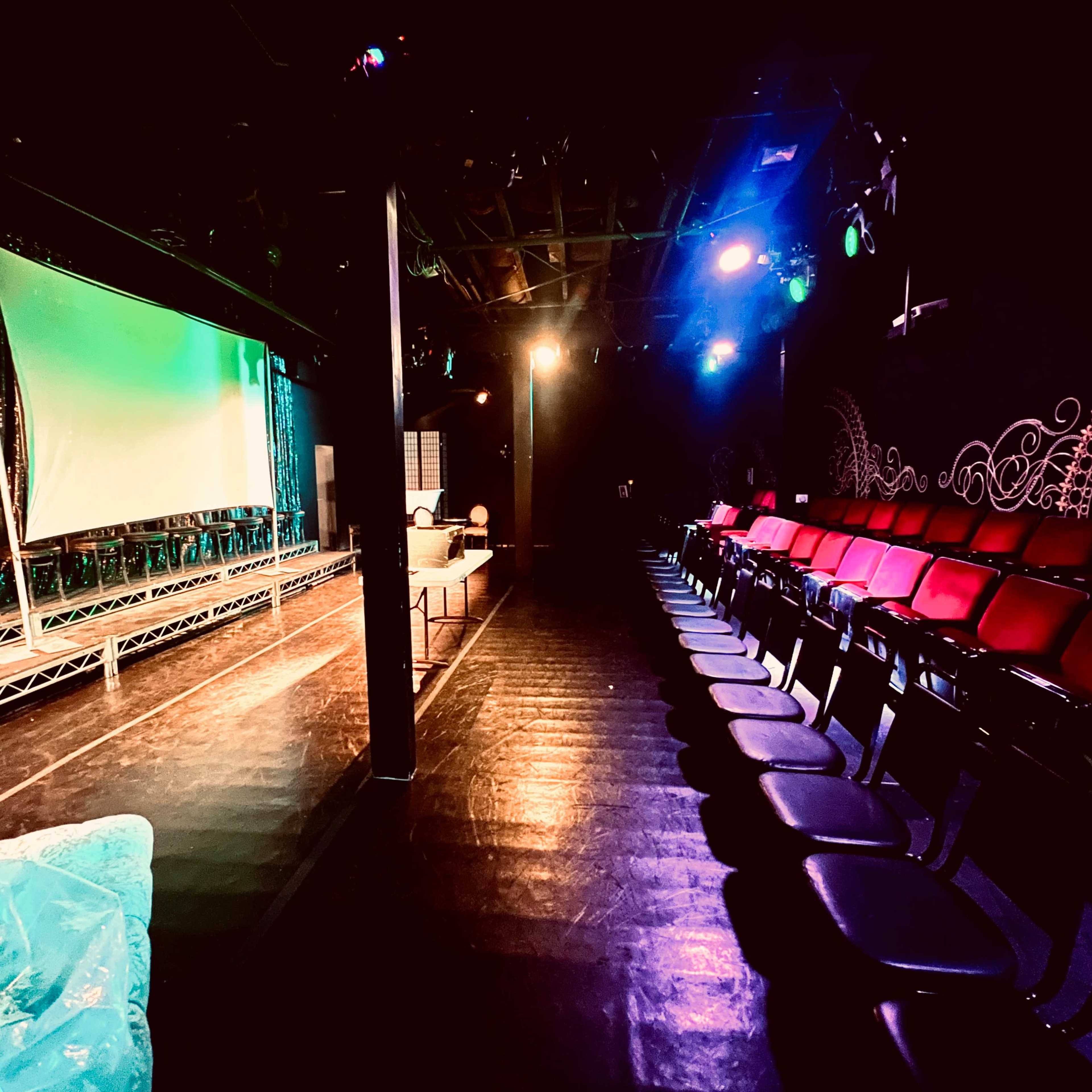 A small theater setting with a stage in the foreground, illuminated by colorful lights, and rows of empty chairs facing the stage.