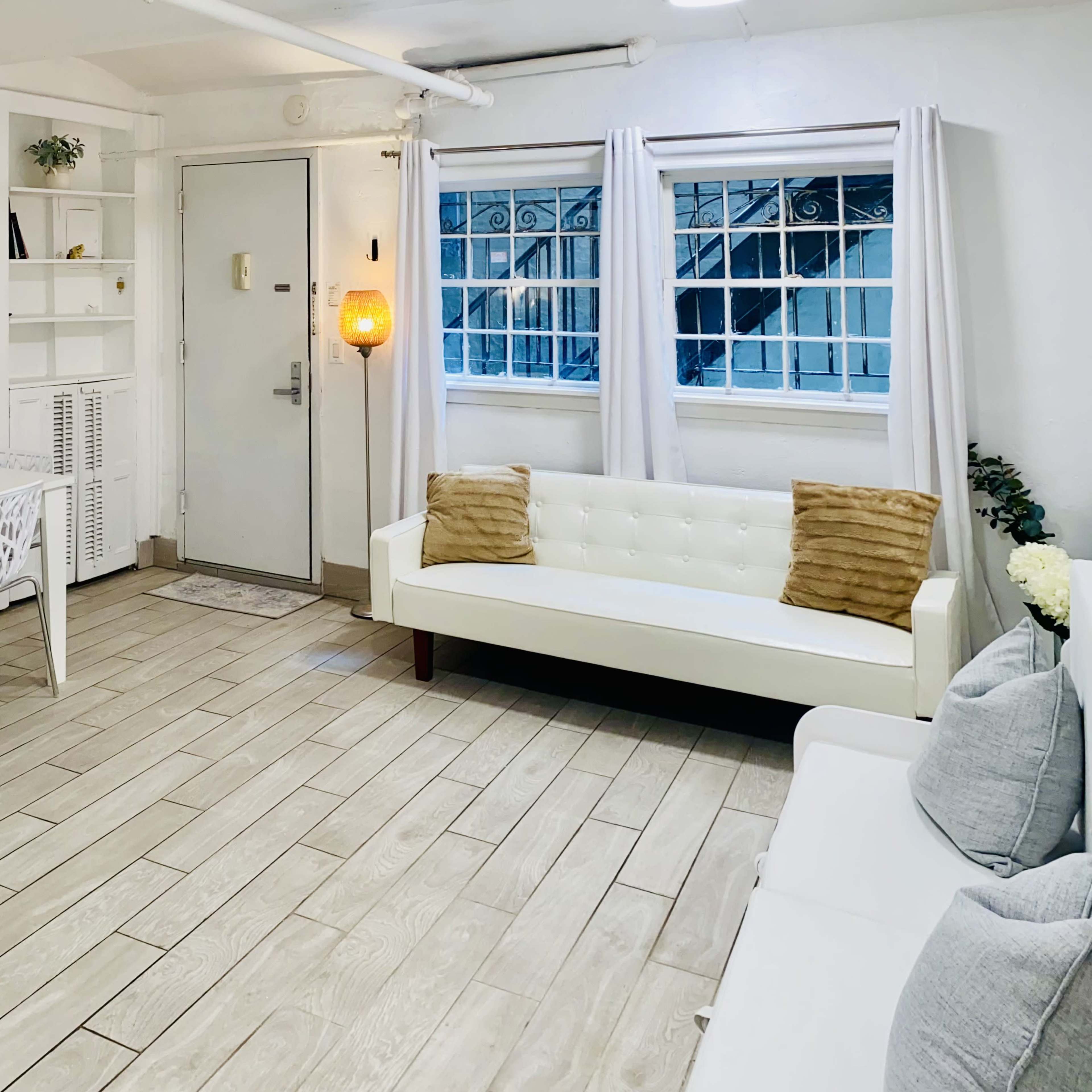 A bright living space features a white sofa, a dining table with chairs, and large windows with sheer curtains.