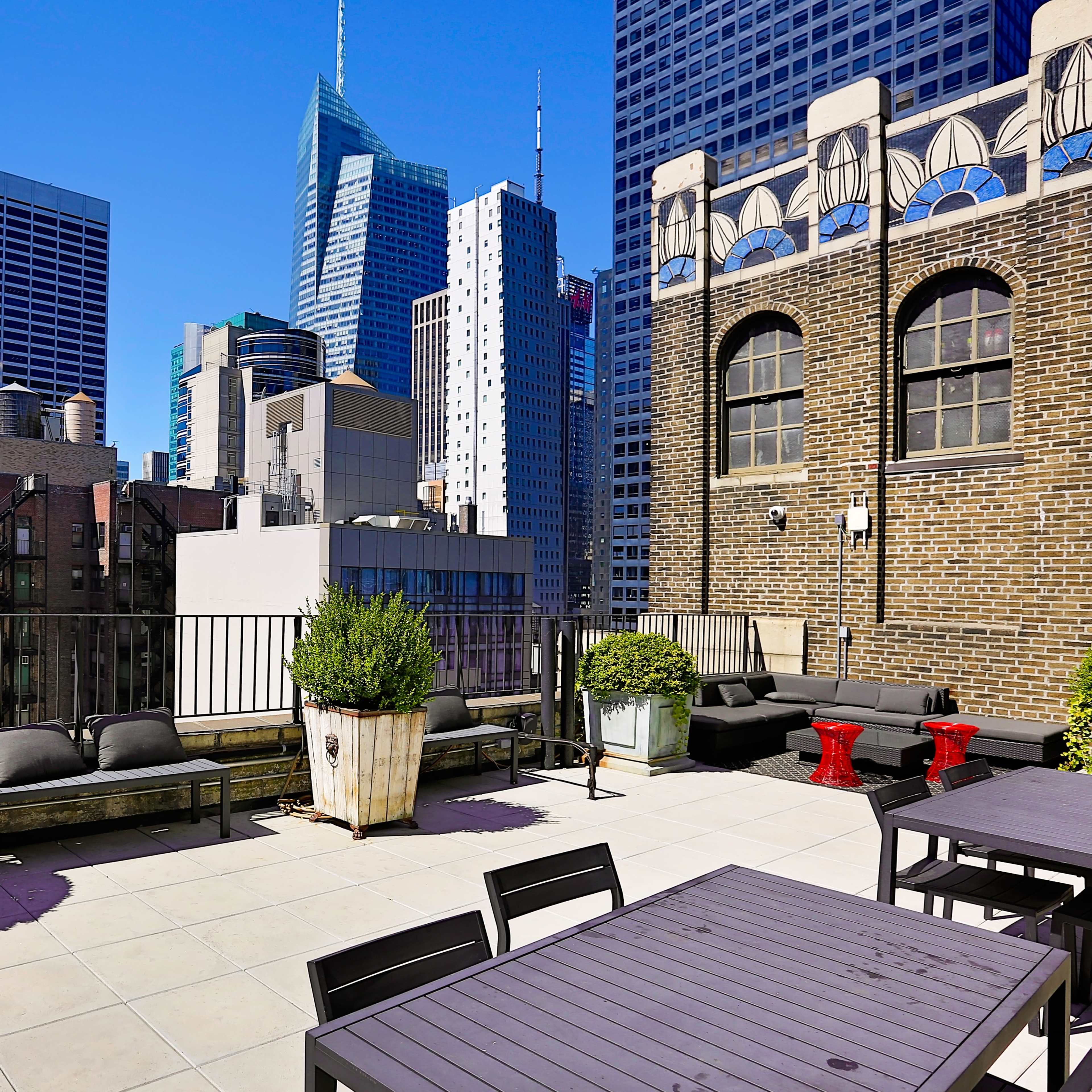 A rooftop terrace features modern seating and potted plants with a clear view of skyscrapers, including the Empire State Building, against a bright blue sky.