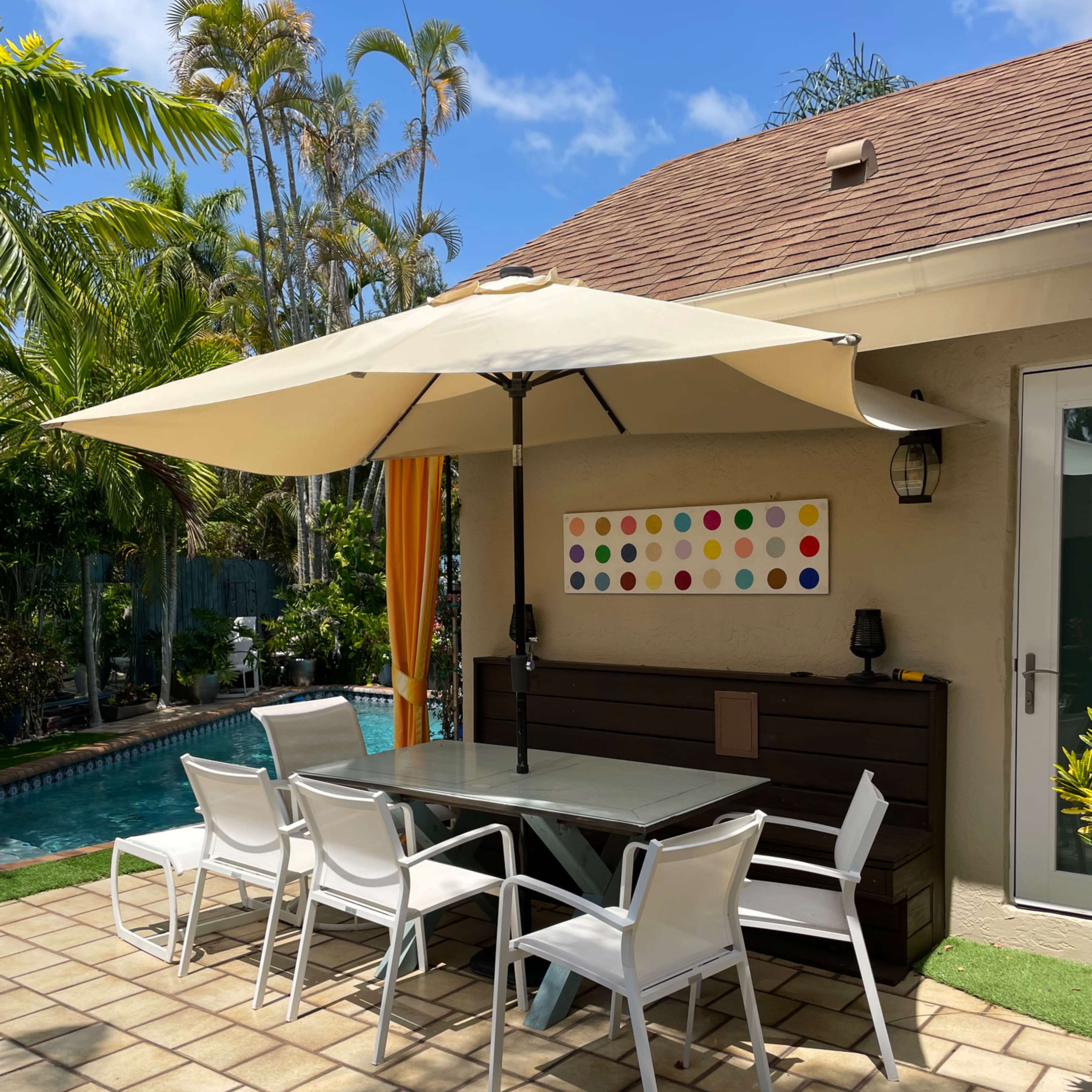A patio area features a large umbrella over a rectangular table surrounded by white chairs, with a pool and palm trees in the background.