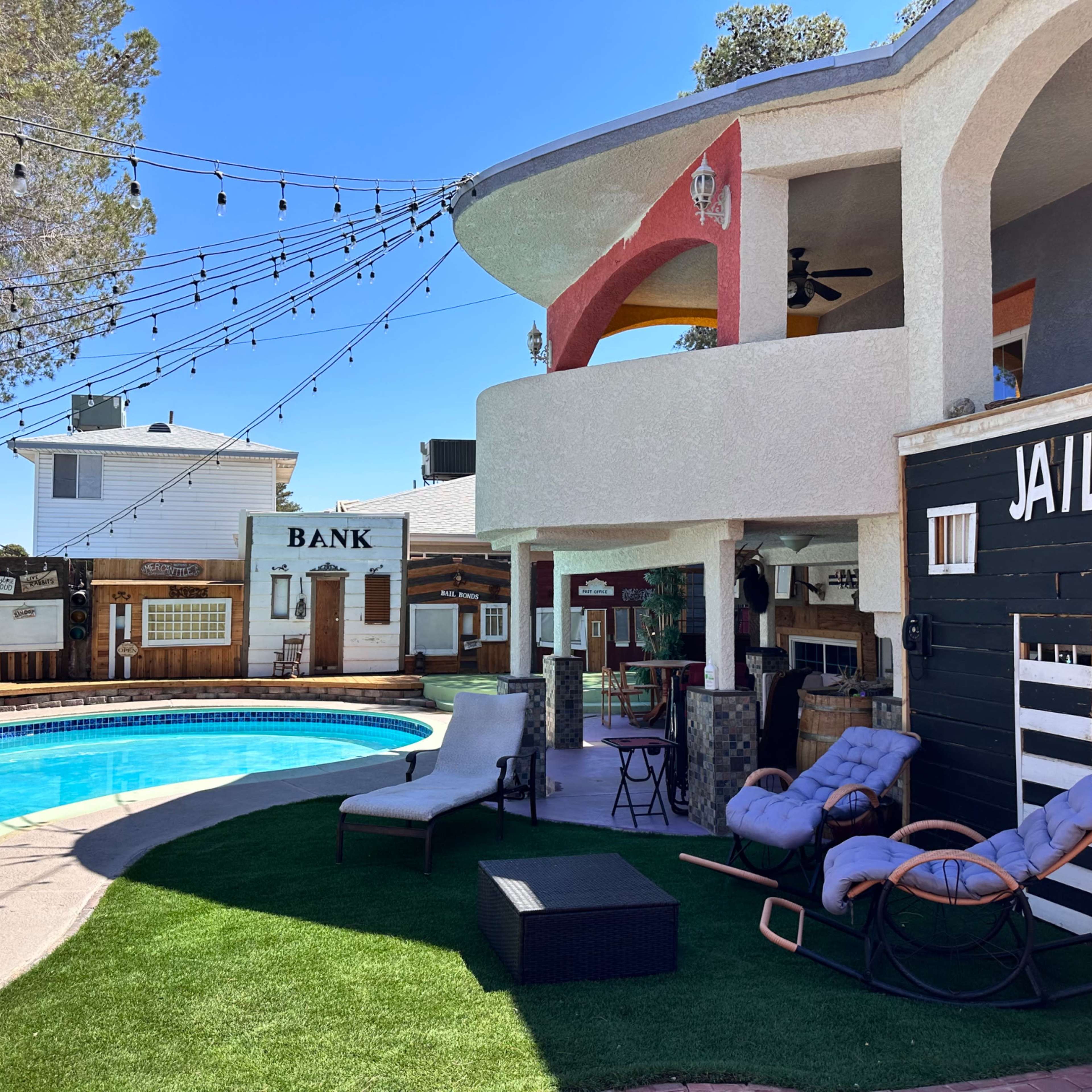The scene features a poolside area with a jail-themed structure and a bank building in the background, surrounded by string lights and lounge chairs.