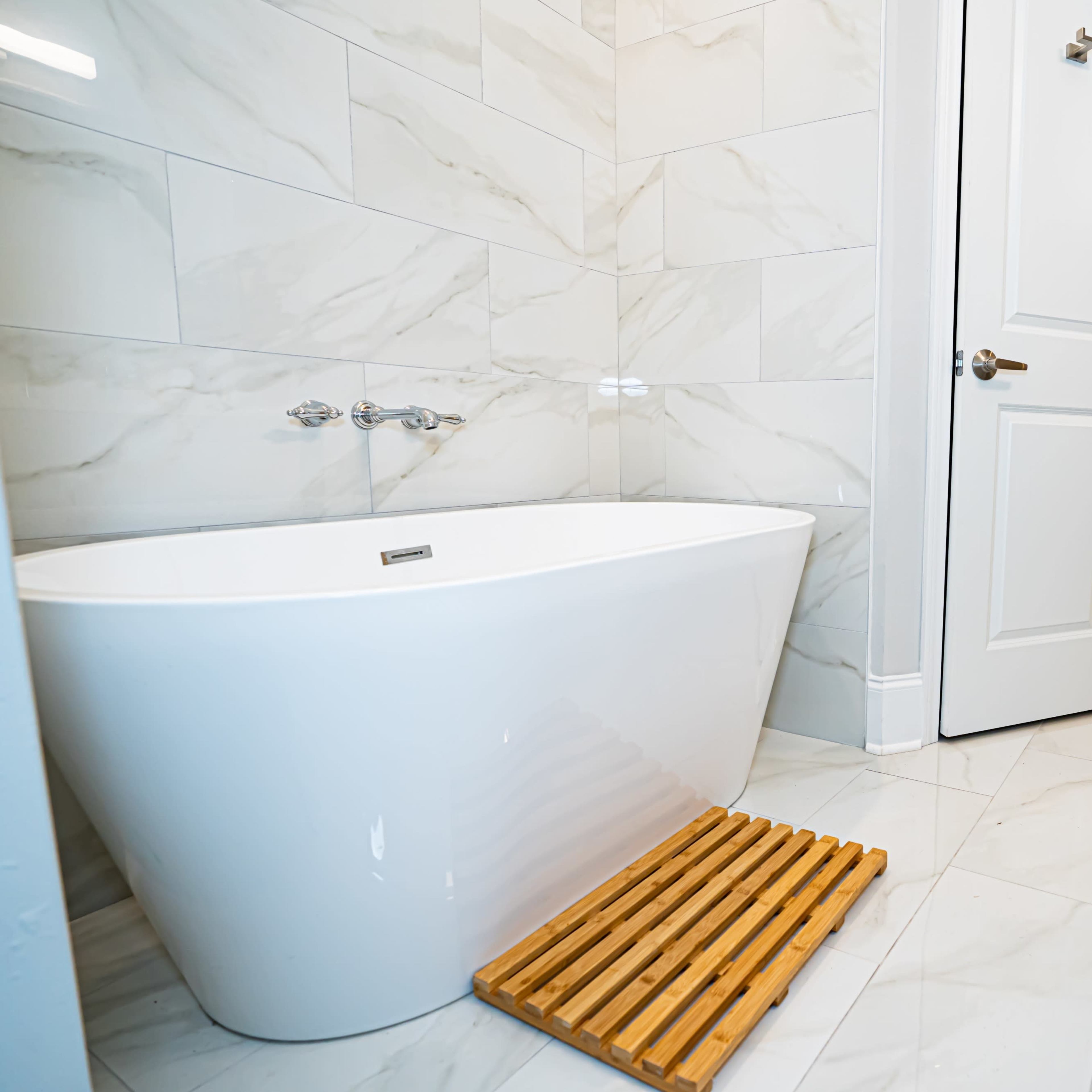A modern bathroom features a freestanding white bathtub next to a wooden bath mat, with marble tile walls and a vanity in the background.