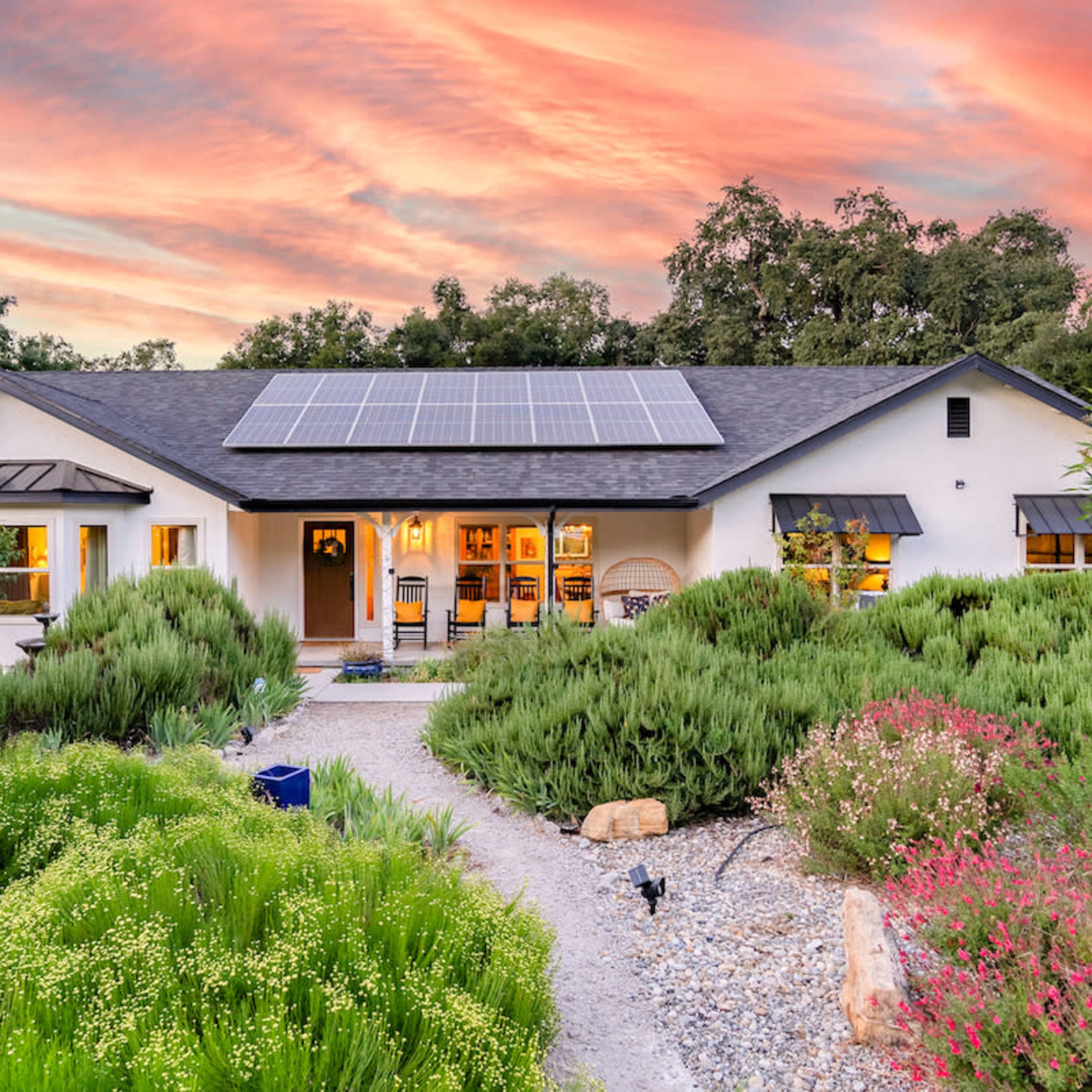 A single-story house with solar panels on the roof is surrounded by a lush, landscaped garden at sunset.
