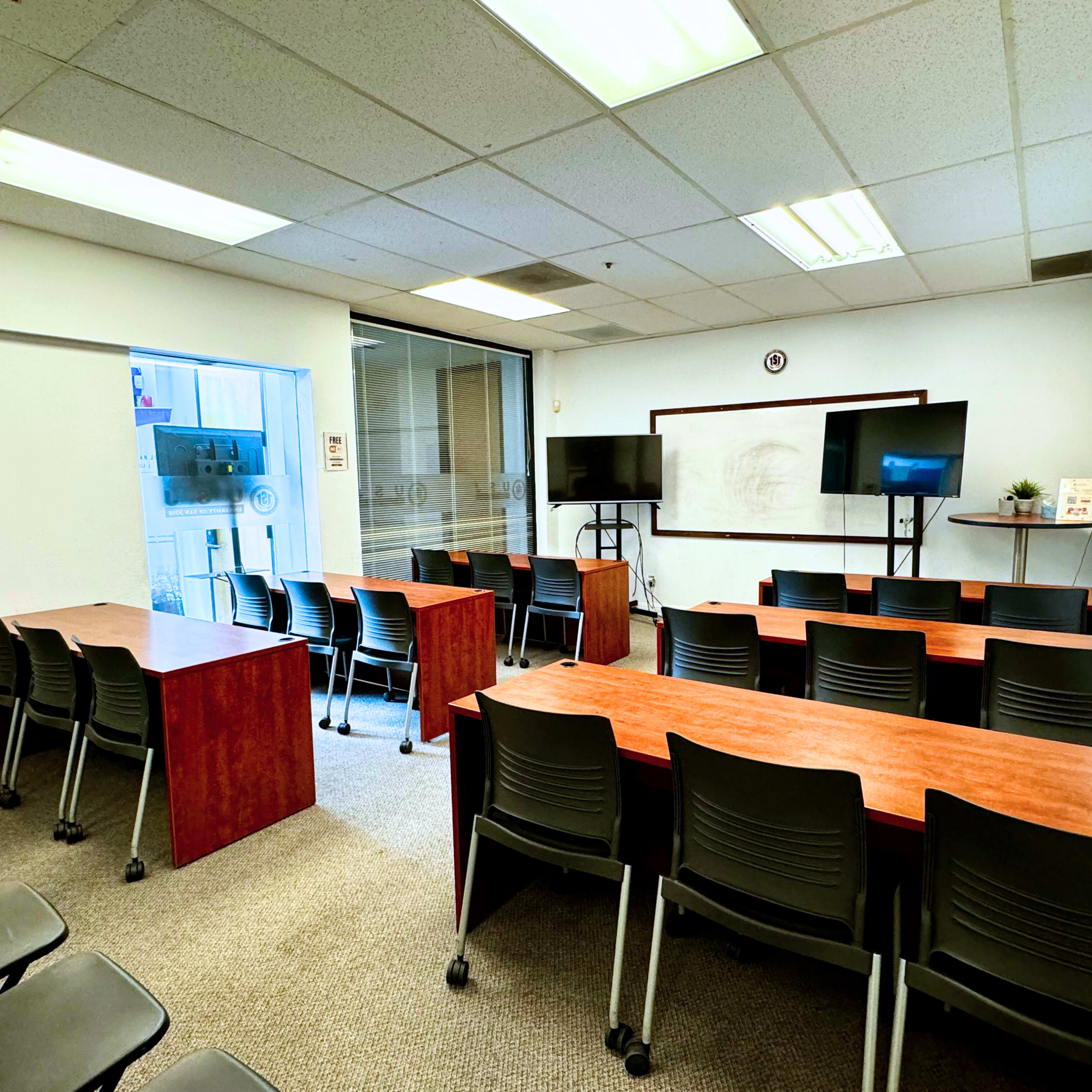 A classroom with rows of tables and chairs, two television screens, and a whiteboard at the front.
