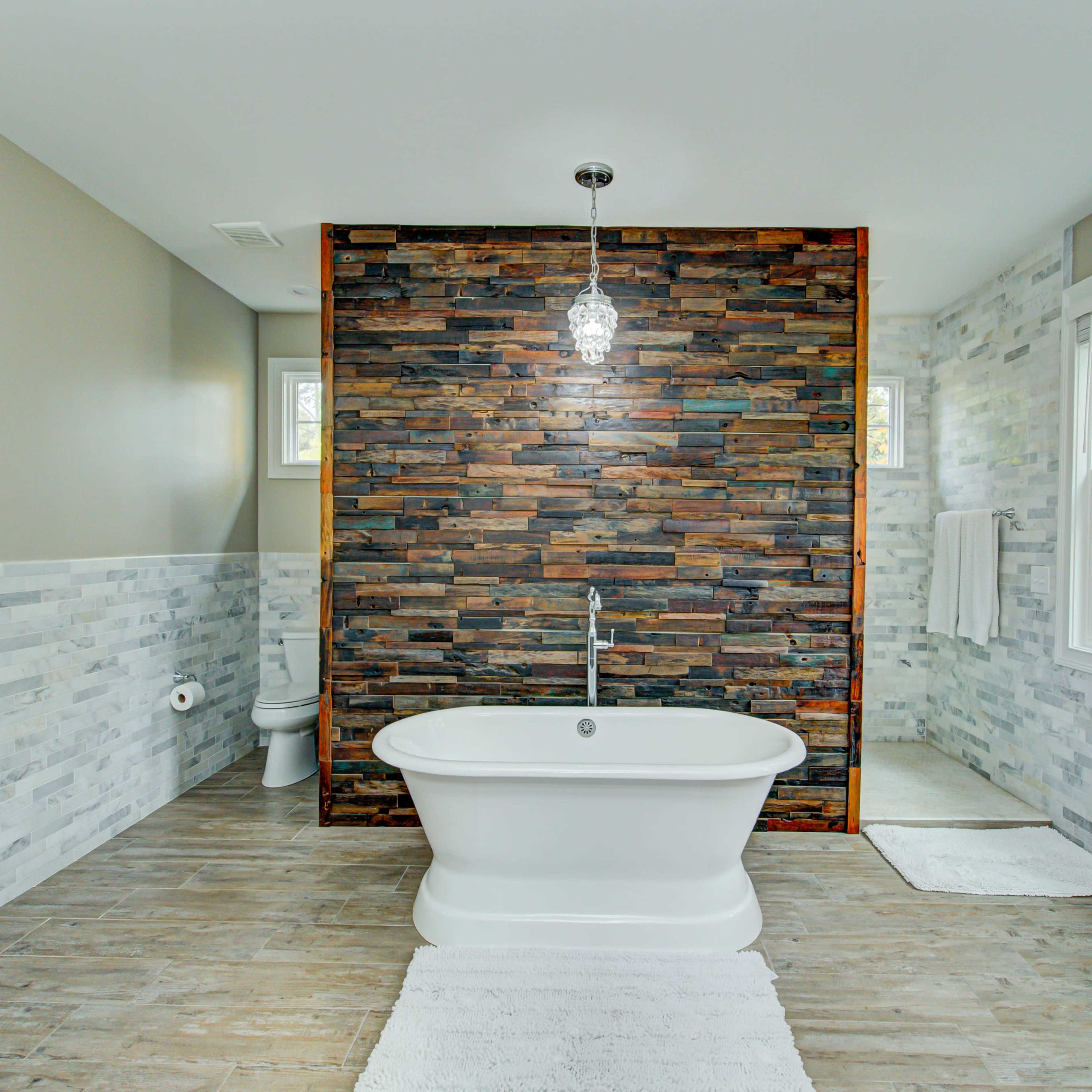 A modern bathroom featuring a freestanding white bathtub, a stone accent wall, and large windows allowing natural light.