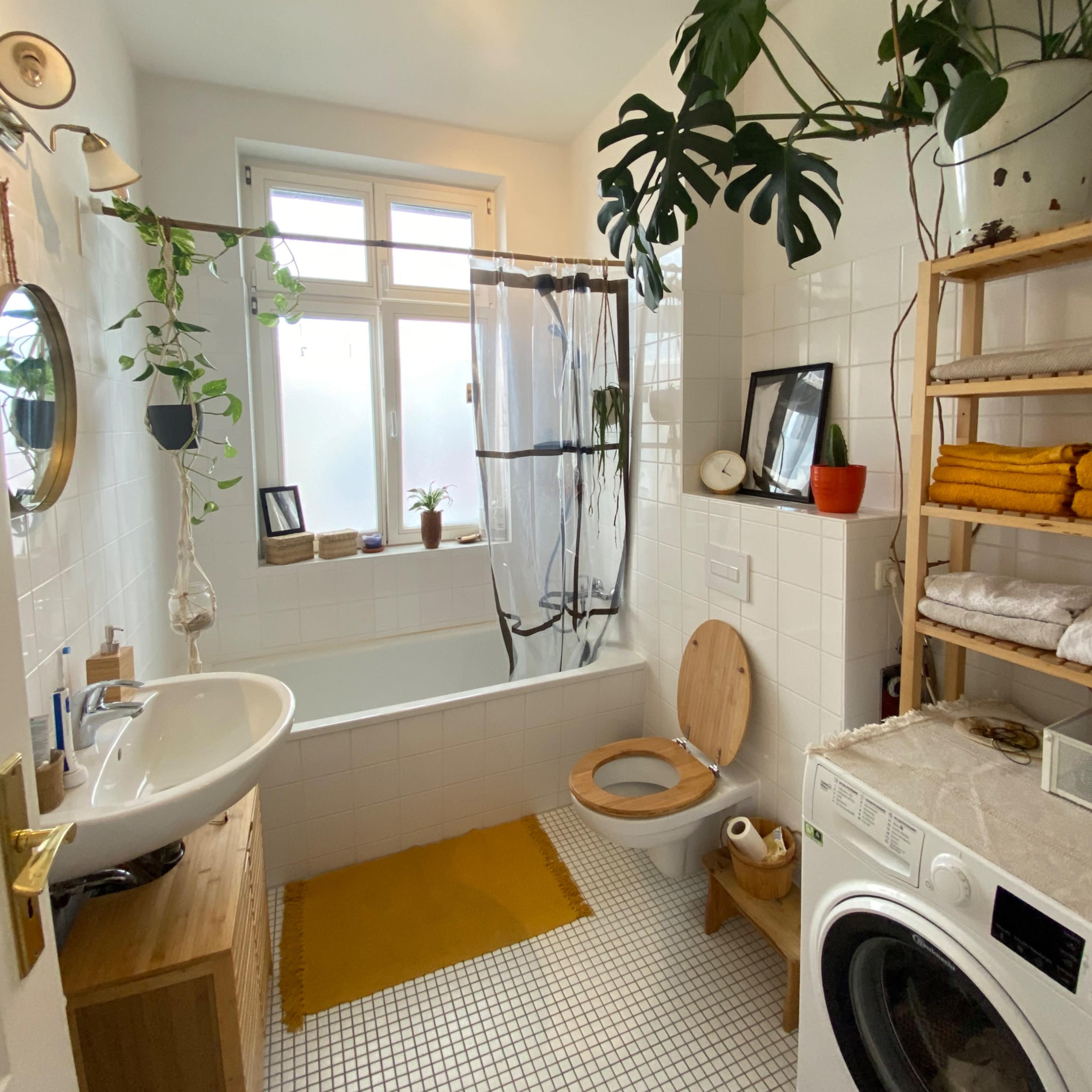 The bathroom features a bathtub with a shower curtain, a wall-mounted sink, a wooden shelf with stacked towels, and a washing machine, all surrounded by plants and natural light.