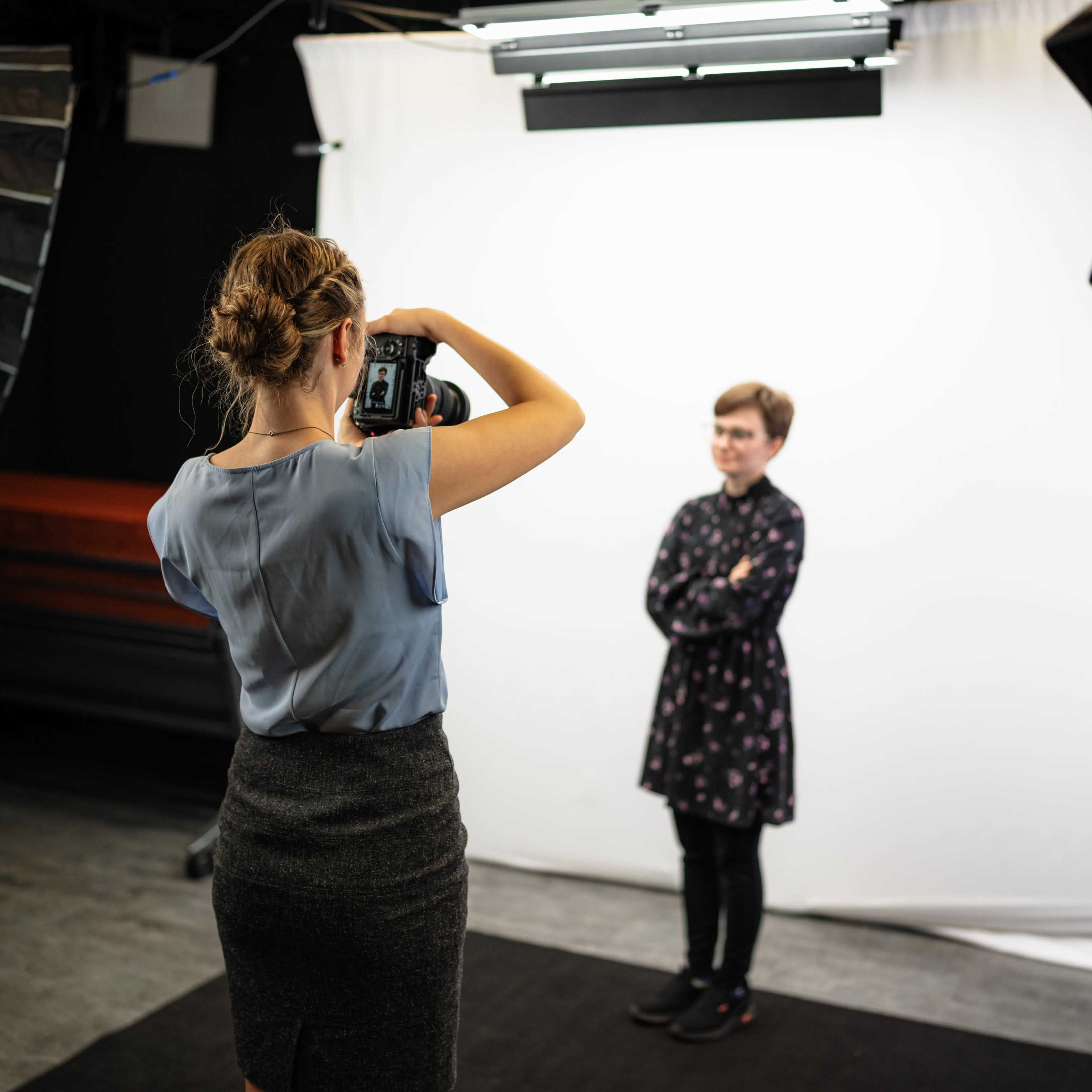 A photographer is taking a portrait of a person standing with crossed arms in front of a white backdrop in a studio.