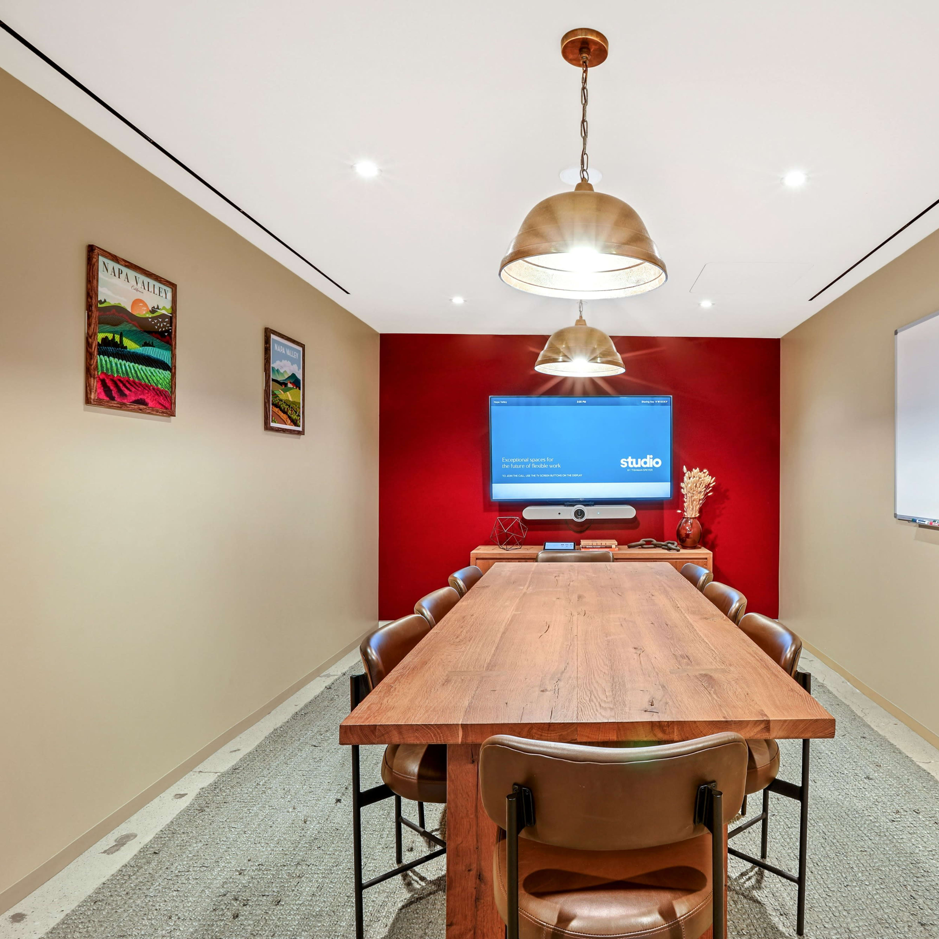 A long wooden conference table is surrounded by brown chairs in a small meeting room featuring a large screen on the wall and a whiteboard.