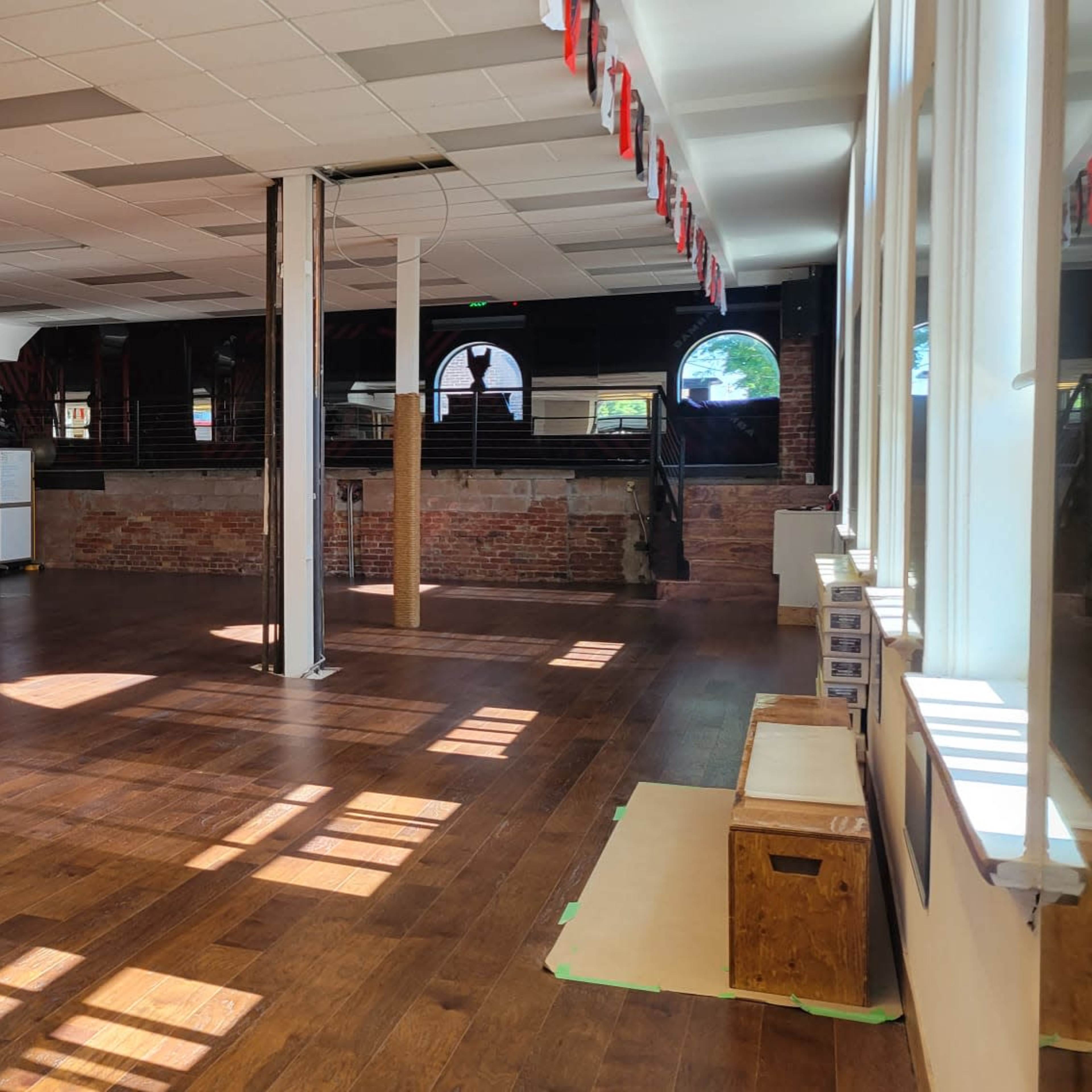 A spacious dance studio with wooden floors, bright natural light streaming in through large windows, and exposed brick walls.