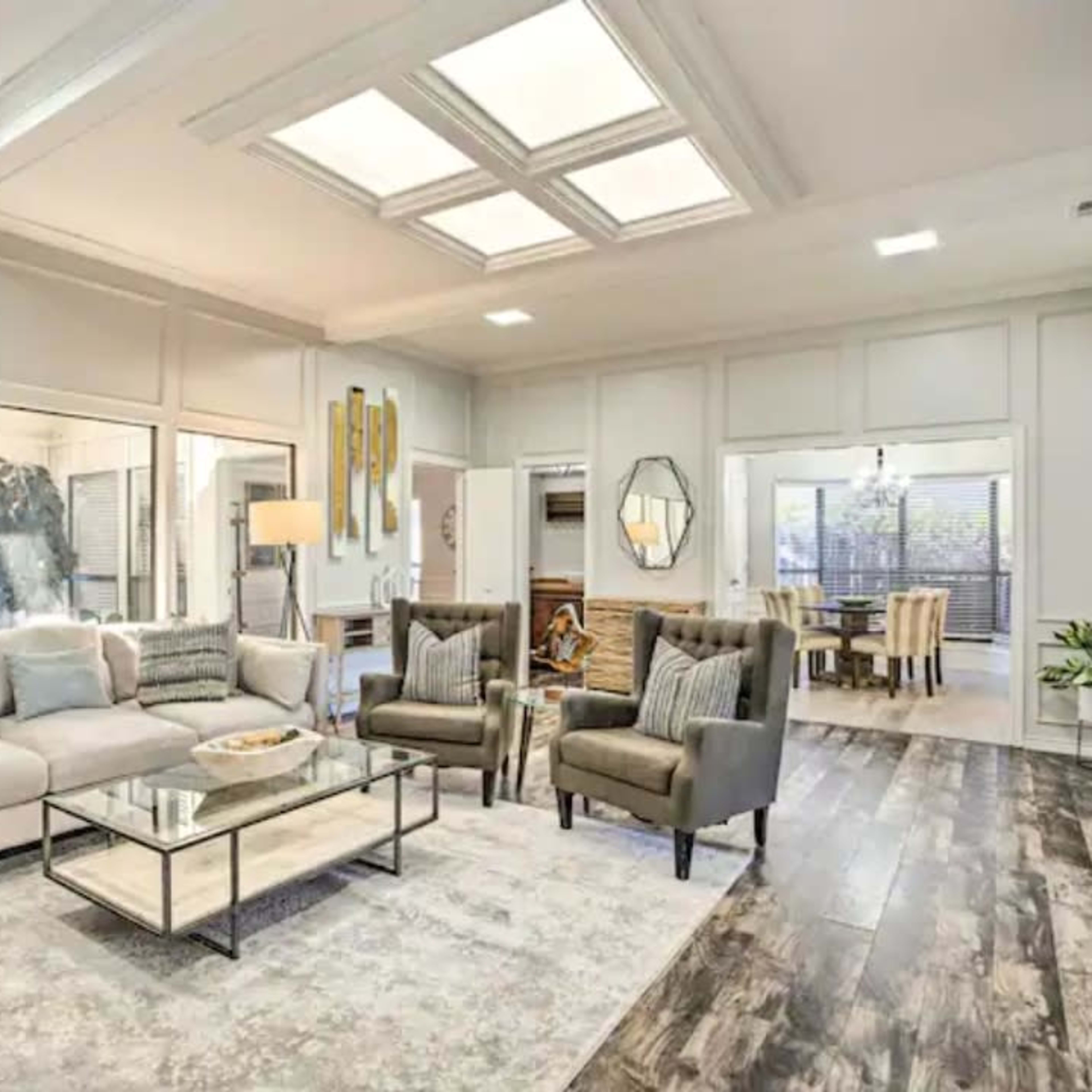 The living room features a light-colored sofa, two armchairs, a glass coffee table, and large windows that overlook a patio area.