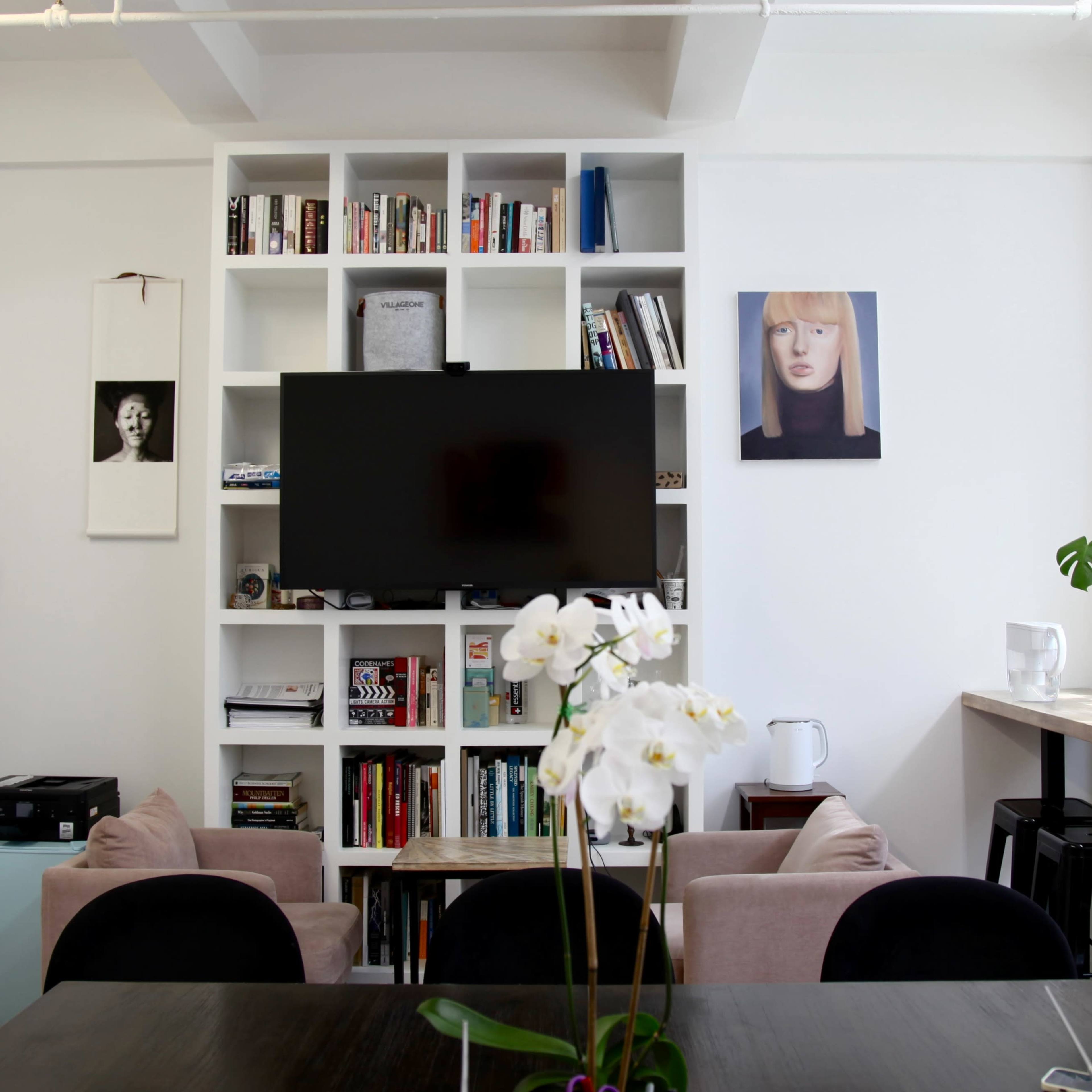 A modern interior features a TV mounted on a white bookshelf filled with books, surrounded by seating and a table with an orchid centerpiece.