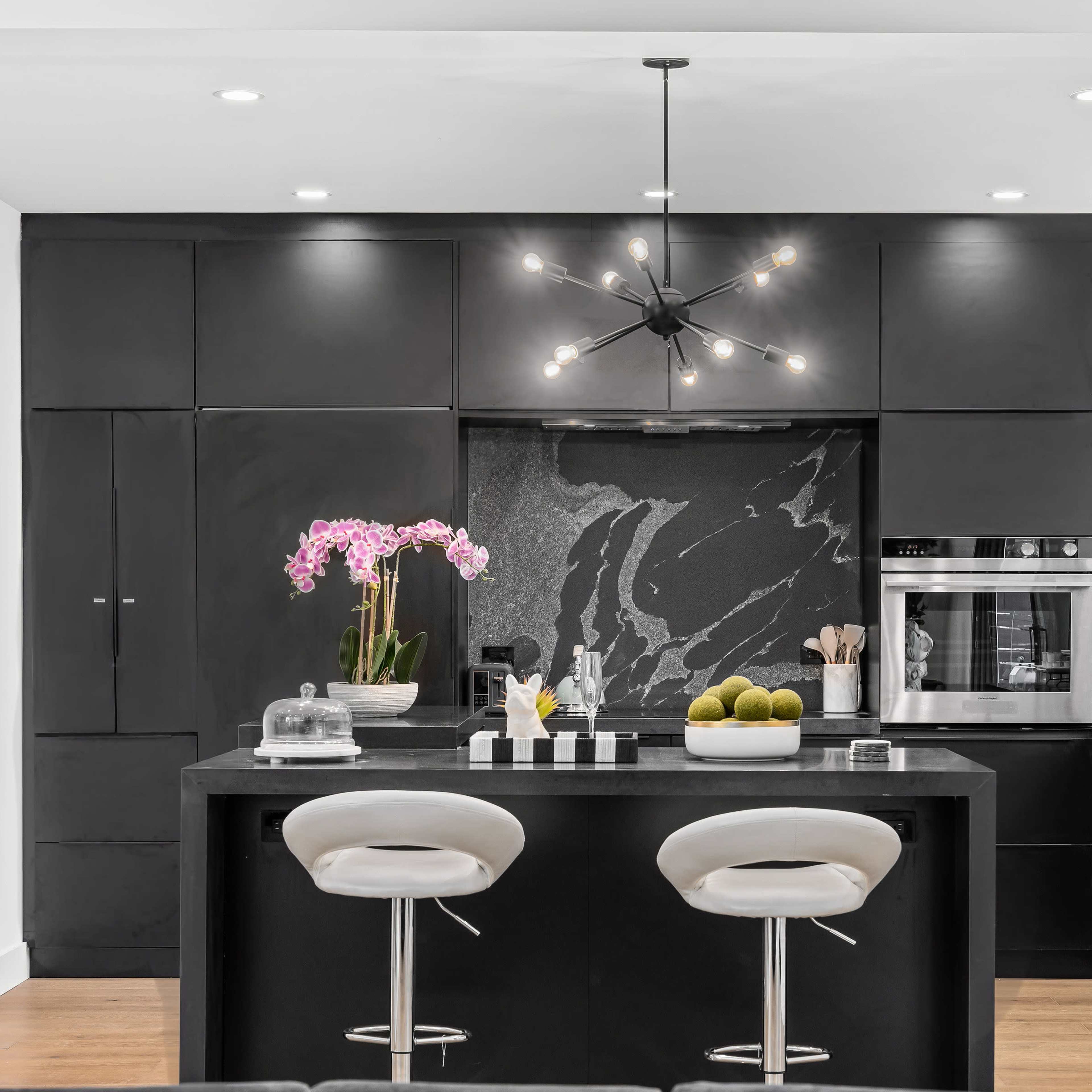 A modern kitchen features black cabinetry, a large island with two white bar stools, and a decorative arrangement of orchids.